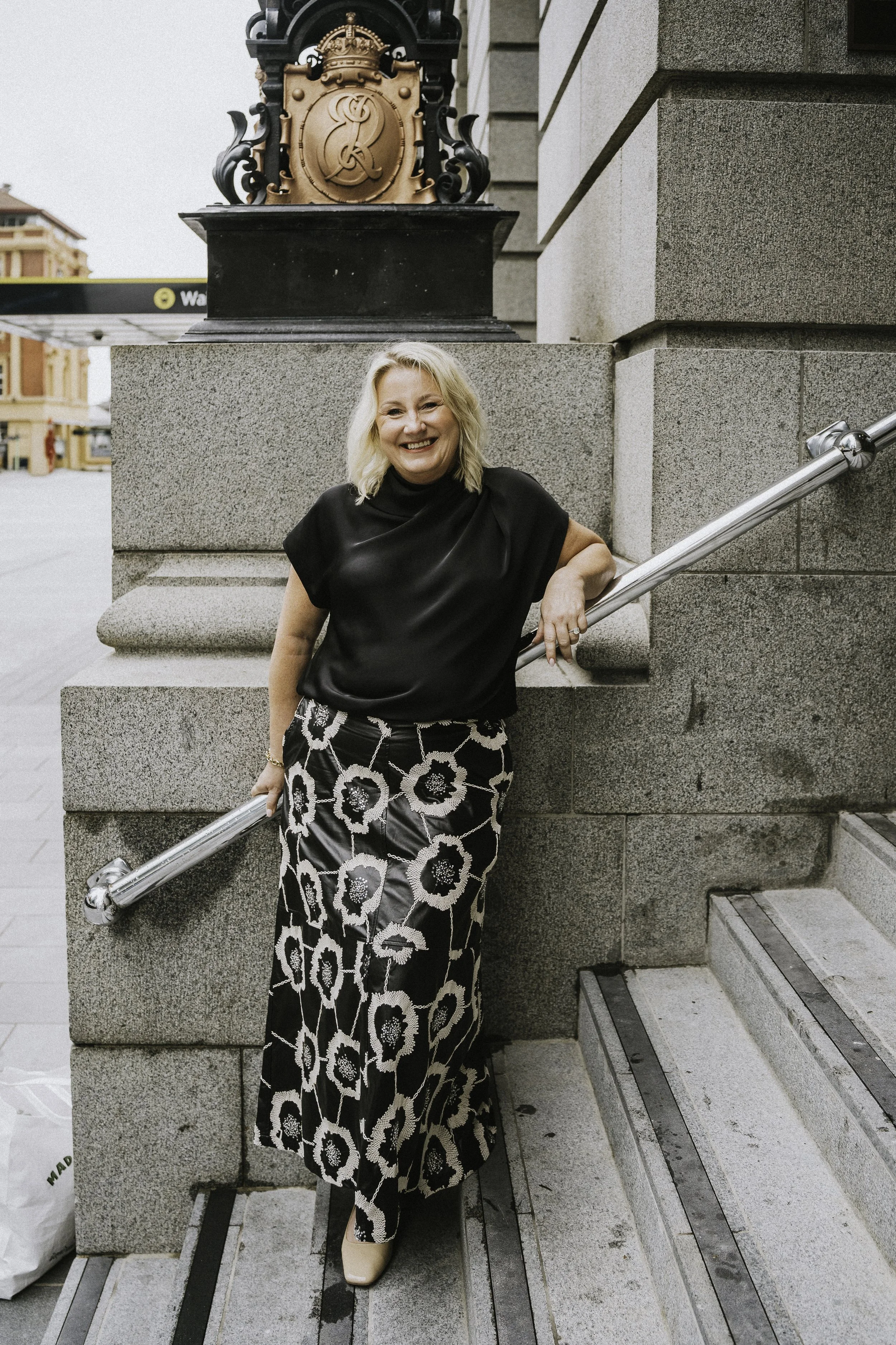 A woman with blonde hair wearing a black top and a long black and white floral skirt, standing on a stairway outside a stone building, smiling at the camera.