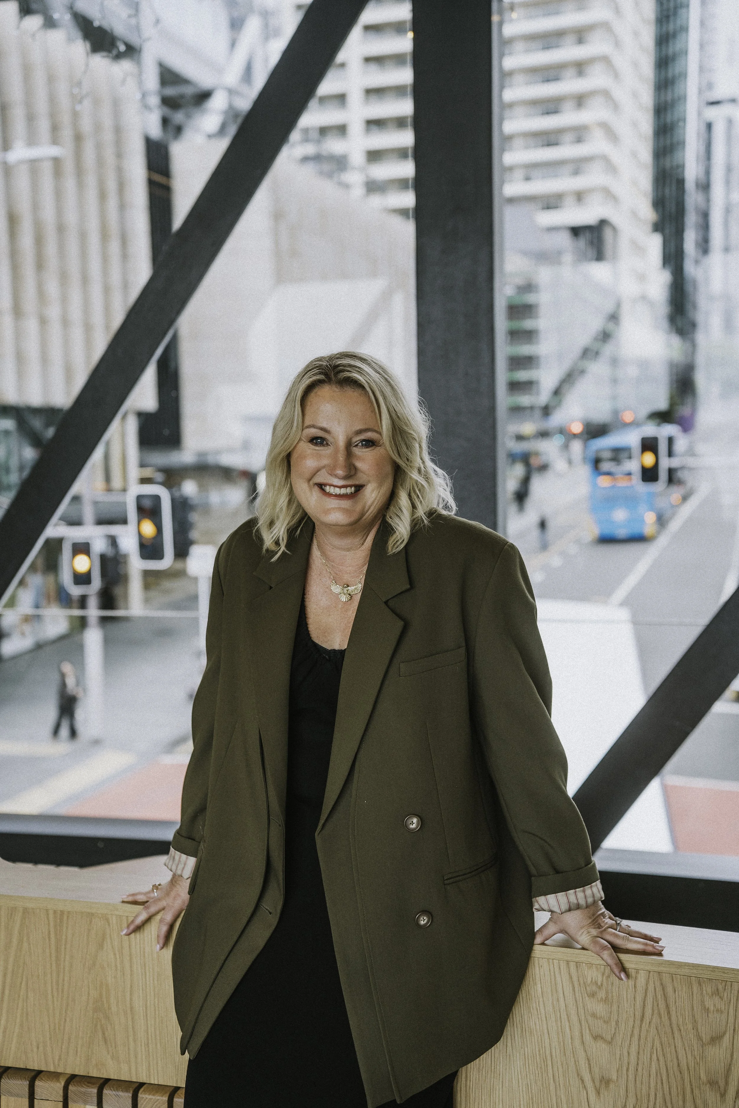 A woman with blonde hair smiling and leaning on a wooden surface, standing in front of a large window overlooking a city street with traffic lights, cars, buses, and high-rise buildings.