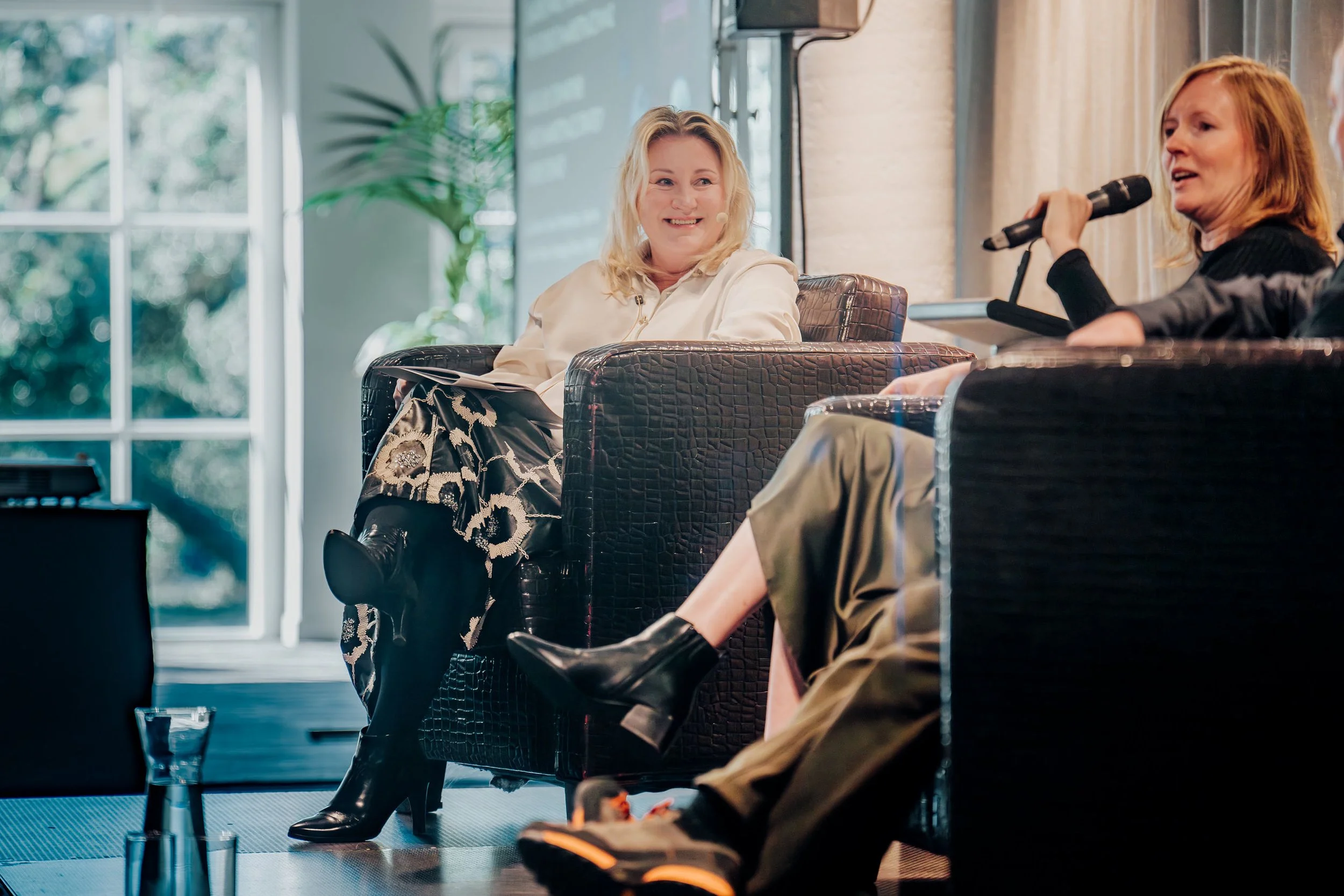 Juanita Neville Te Rito  seated and conversing during a panel discussion or interview on a stage, one holding a microphone, with large windows and a plant in the background.