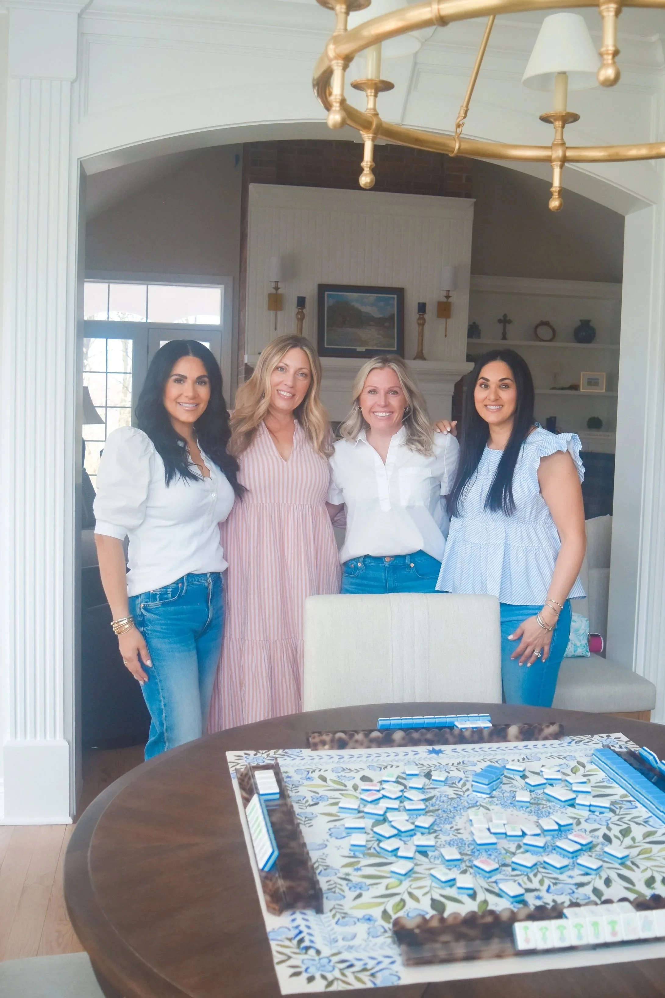 Four women standing around a dining table with a board game, smiling in a bright, home interior with a fireplace and window in the background.