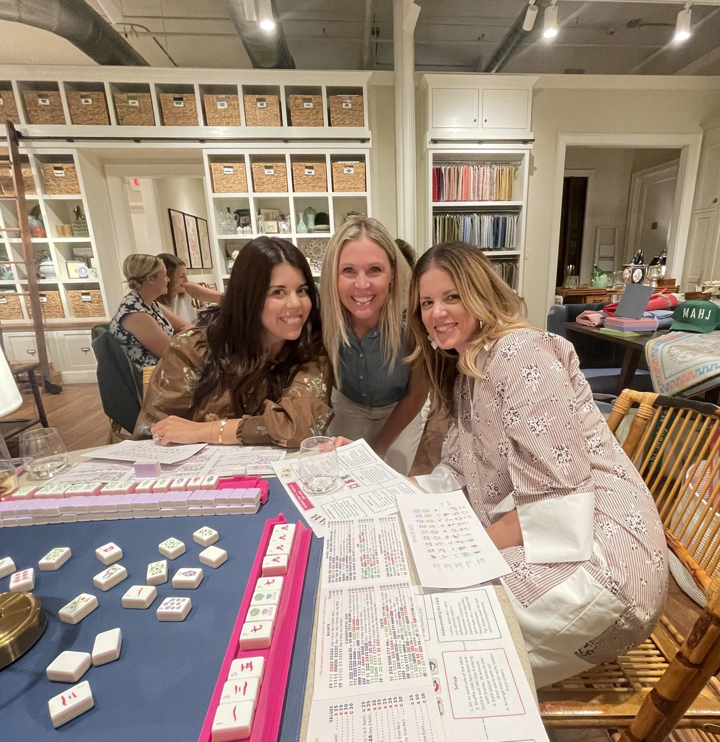 Three women smiling and leaning over a table playing Mahjong in a cozy, well-lit room with shelves of books and wicker baskets in the background.