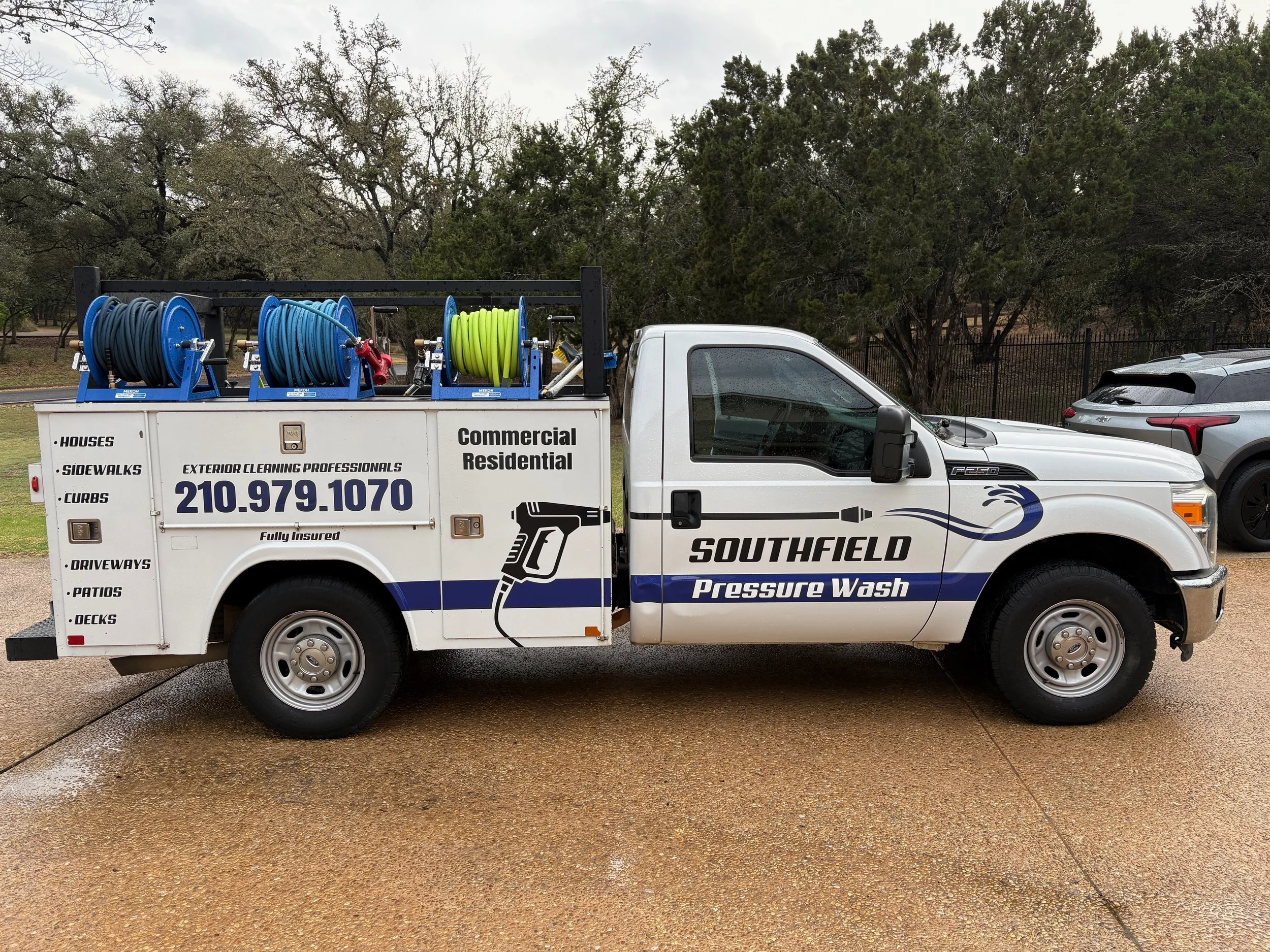 White service truck with hoses on top, labeled 'Southfield Pressure Wash,' parked on a wet concrete surface with trees in the background.