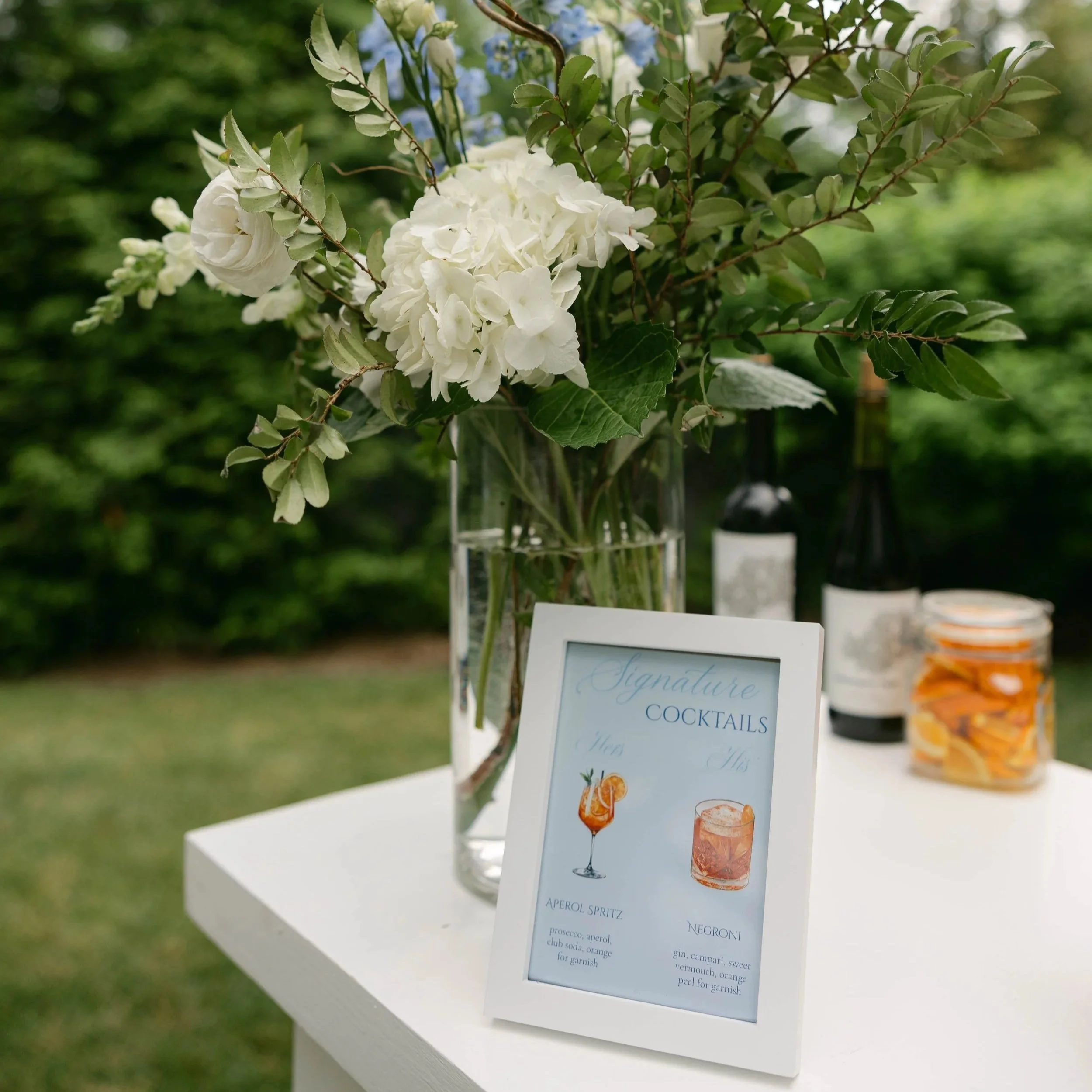 A close-up of a sign with a cocktail menu featuring drinks like Aperol Spritz and Negroni, with a bouquet of white flowers and greenery in the background.