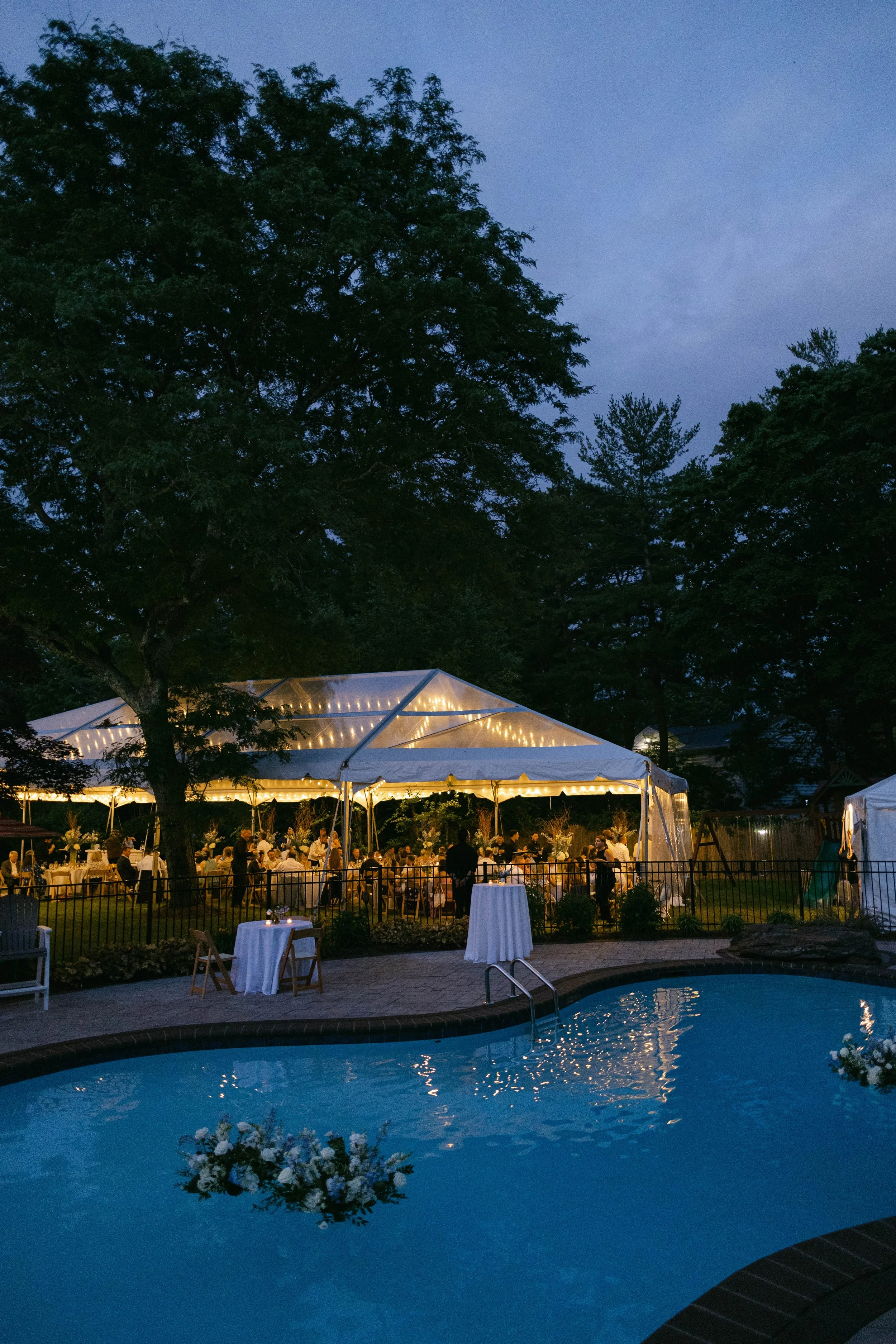 An outdoor evening event with a large tent illuminated by string lights, surrounded by tables and chairs with guests. There is a swimming pool in the foreground with reflections and flowers near the water's edge, and tall trees in the background unde