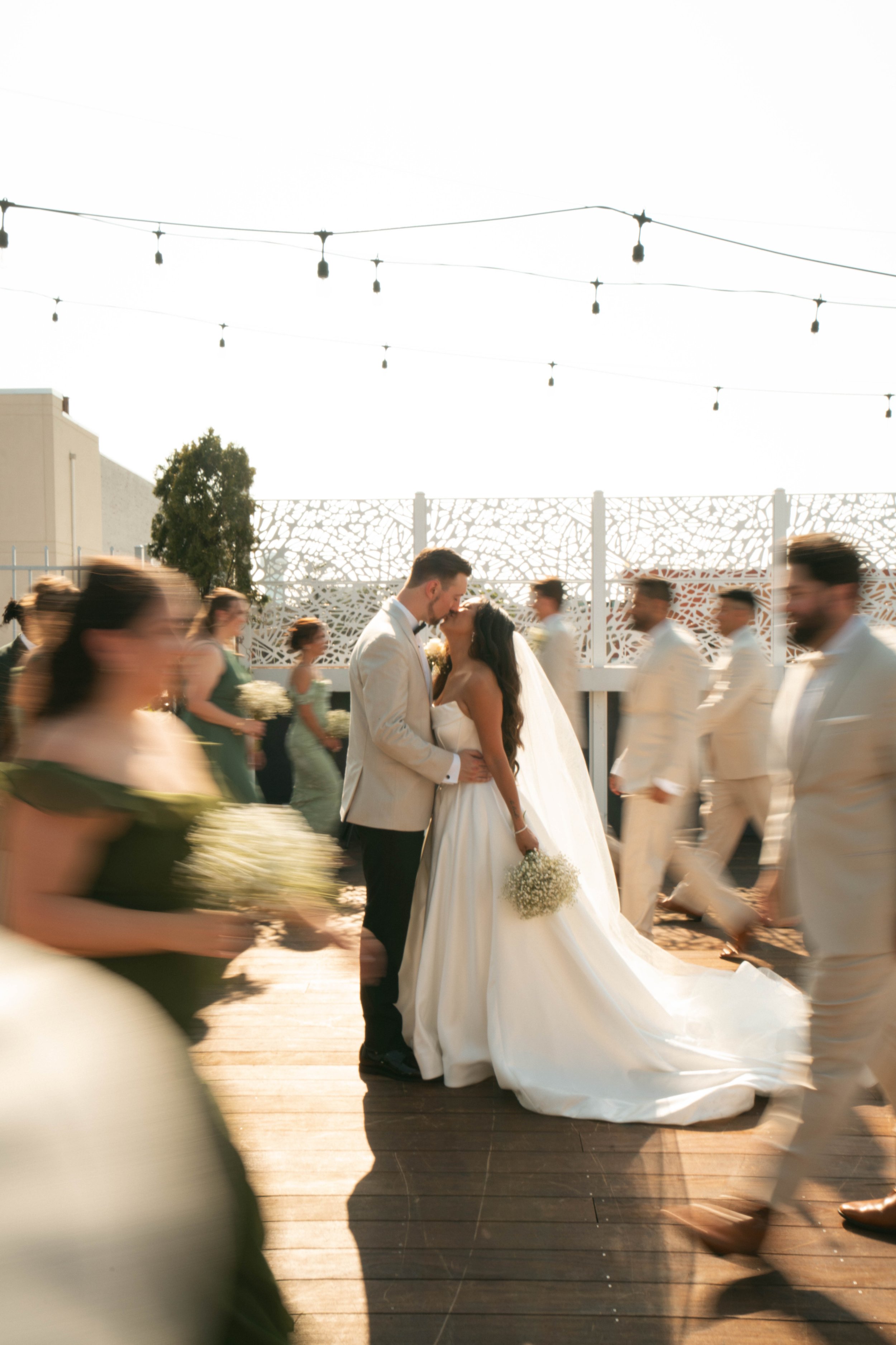 A bride and groom kiss during their wedding reception as they dance on a wooden floor, surrounded by blurred wedding guests in motion. The bride wears a white strapless gown, holds a bouquet of baby's breath, and the groom wears a light-colored suit 