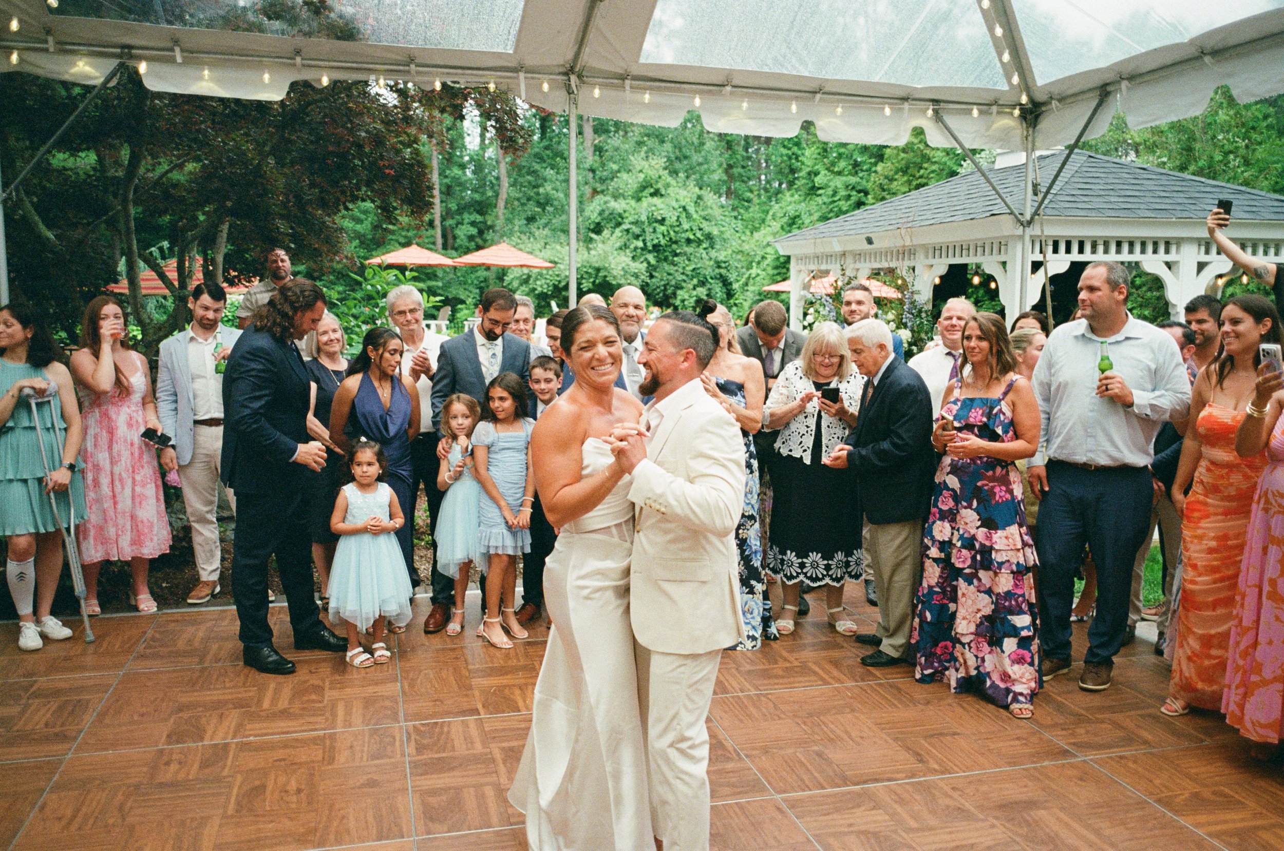 A newlywed couple sharing their first dance at an outdoor wedding reception, surrounded by family and friends.