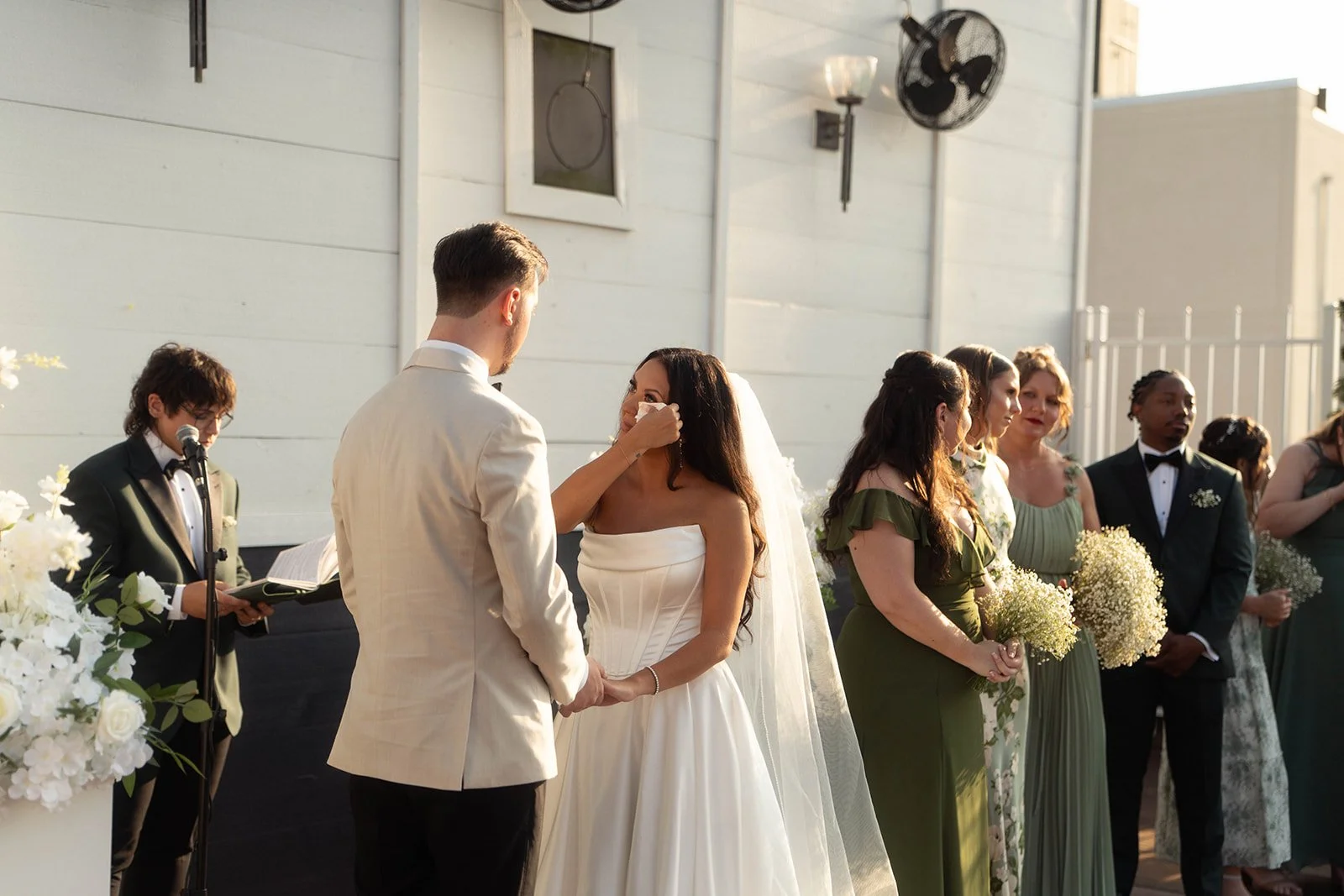 A wedding ceremony outdoors with a bride and groom holding hands, surrounded by bridesmaids and groomsmen. The groom is wiping the bride's face with a tissue. The officiant is reading from a book, and there are floral arrangements and a decorated bac