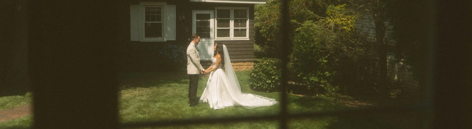 A bride and groom holding hands and facing each other in a yard, seen through a window with blinds. The bride is in a white wedding gown with a long train and veil, and the groom is in a beige suit with black pants.