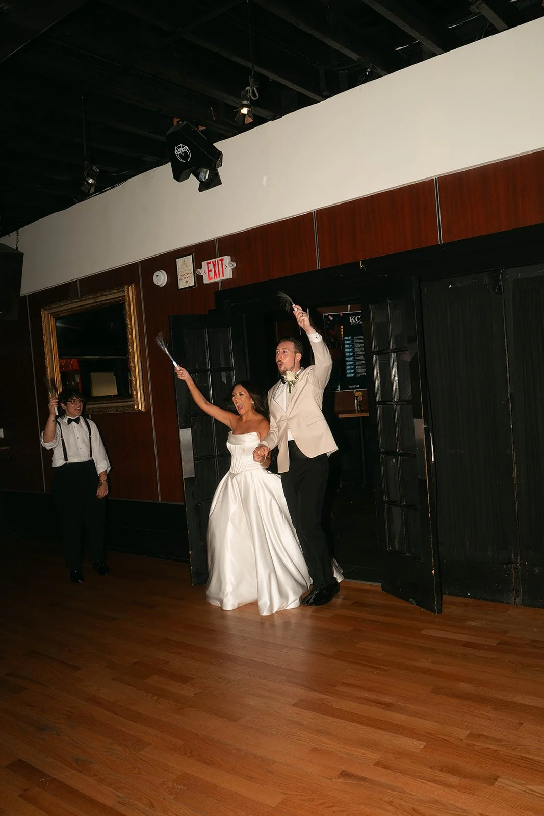A newlywed couple celebrating at their wedding reception, walking out of a door while holding hands, with the bride in a white wedding gown and the groom in a white tuxedo jacket, as they toss confetti or flower petals.
