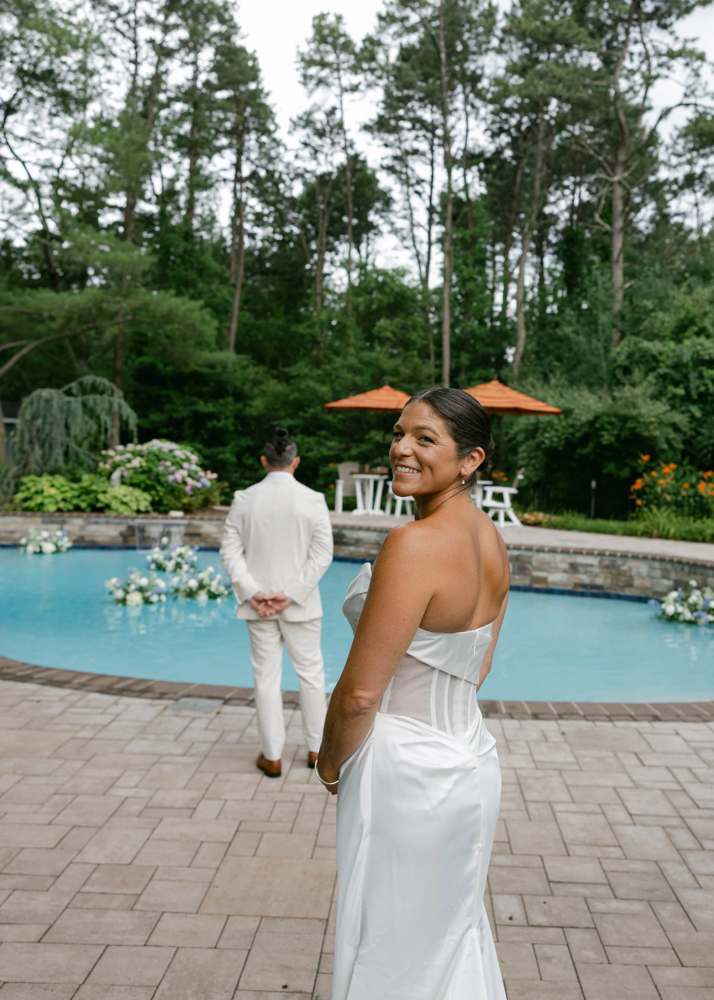 A smiling woman in a white dress stands by a pool with a man in a white suit in the background, near patio furniture and orange umbrellas. There are trees and greenery surrounding the area, indicating an outdoor setting.