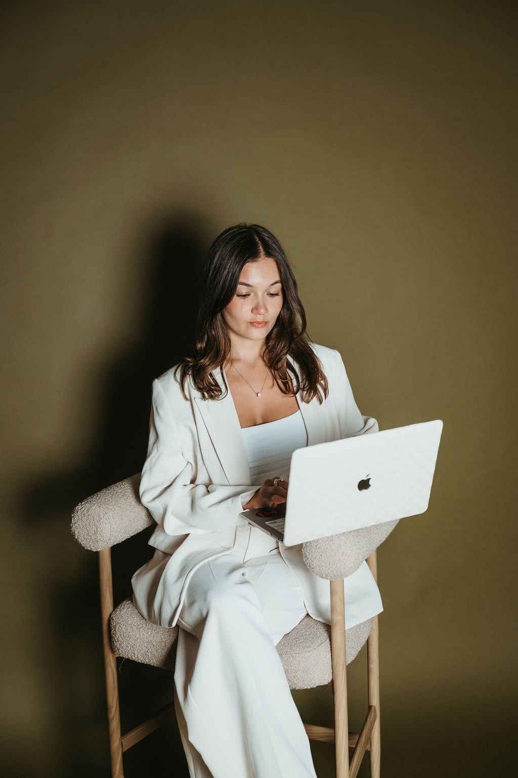 A young woman with dark hair, wearing a white suit, sitting on a chair while working on a white Apple MacBook laptop against a plain olive green wall.