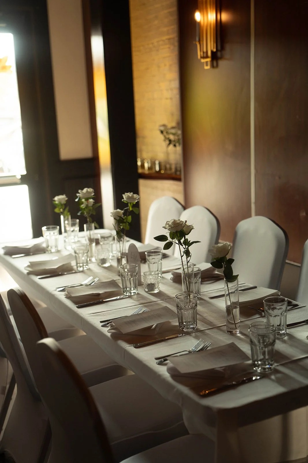 Elegant dining table set for a formal event with white roses in tall vases, water glasses, napkins, and silverware, in a warmly lit room.