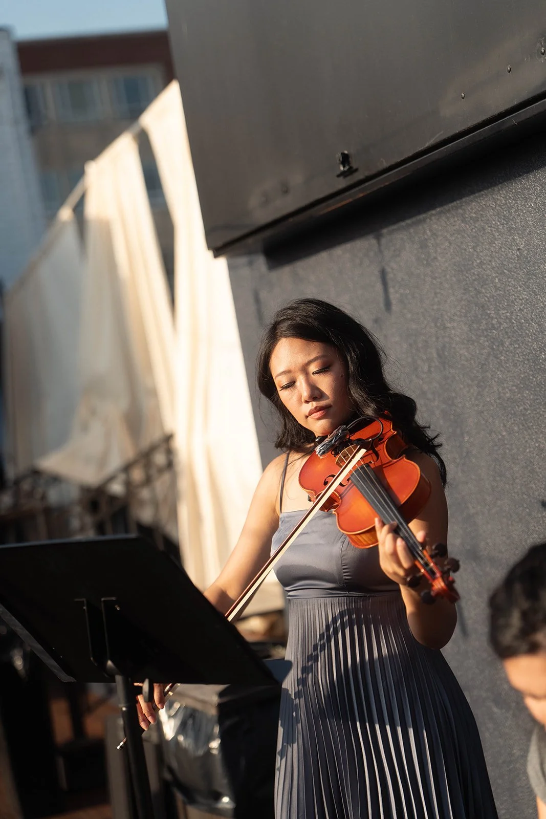 A woman playing a violin outdoors. She is wearing a sleeveless, pleated gray dress, with a music stand in front of her, and appears to be performing.