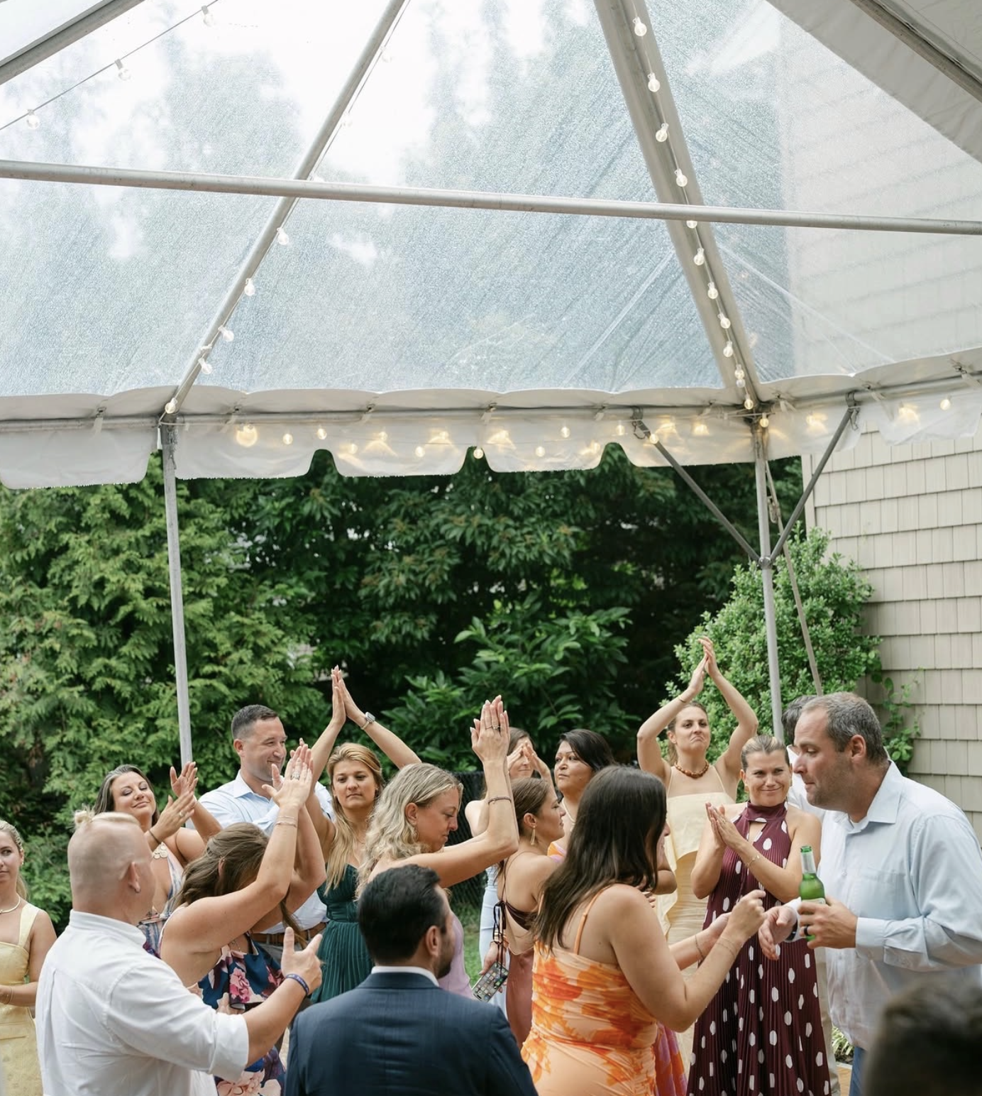 Group of people dancing and socializing under a large outdoor tent with string lights during daytime.