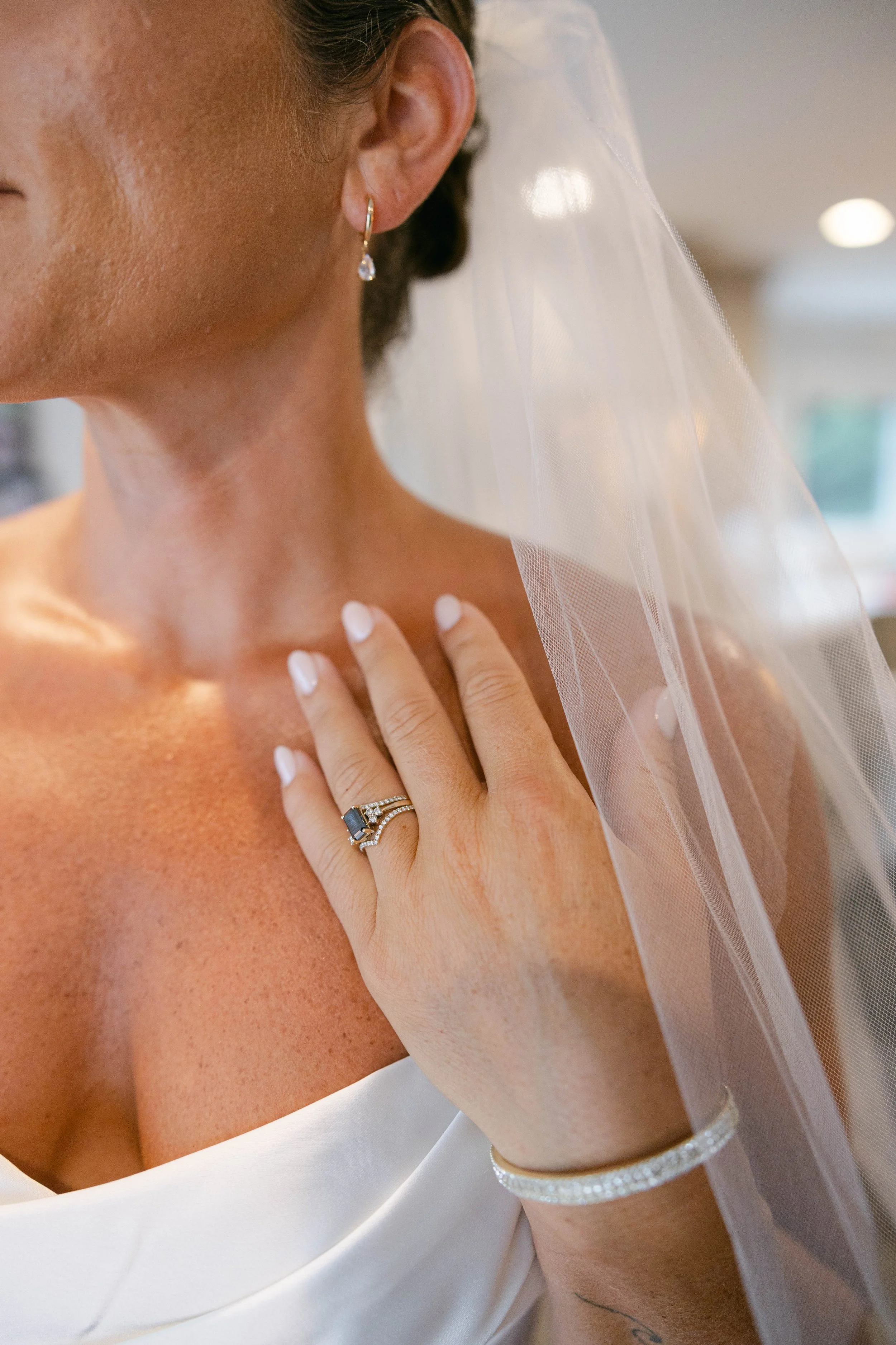 A close-up of a bride showing her neck, shoulder, and hand, wearing a white dress, a diamond ring, diamond bracelet, and diamond earrings, with a sheer veil.