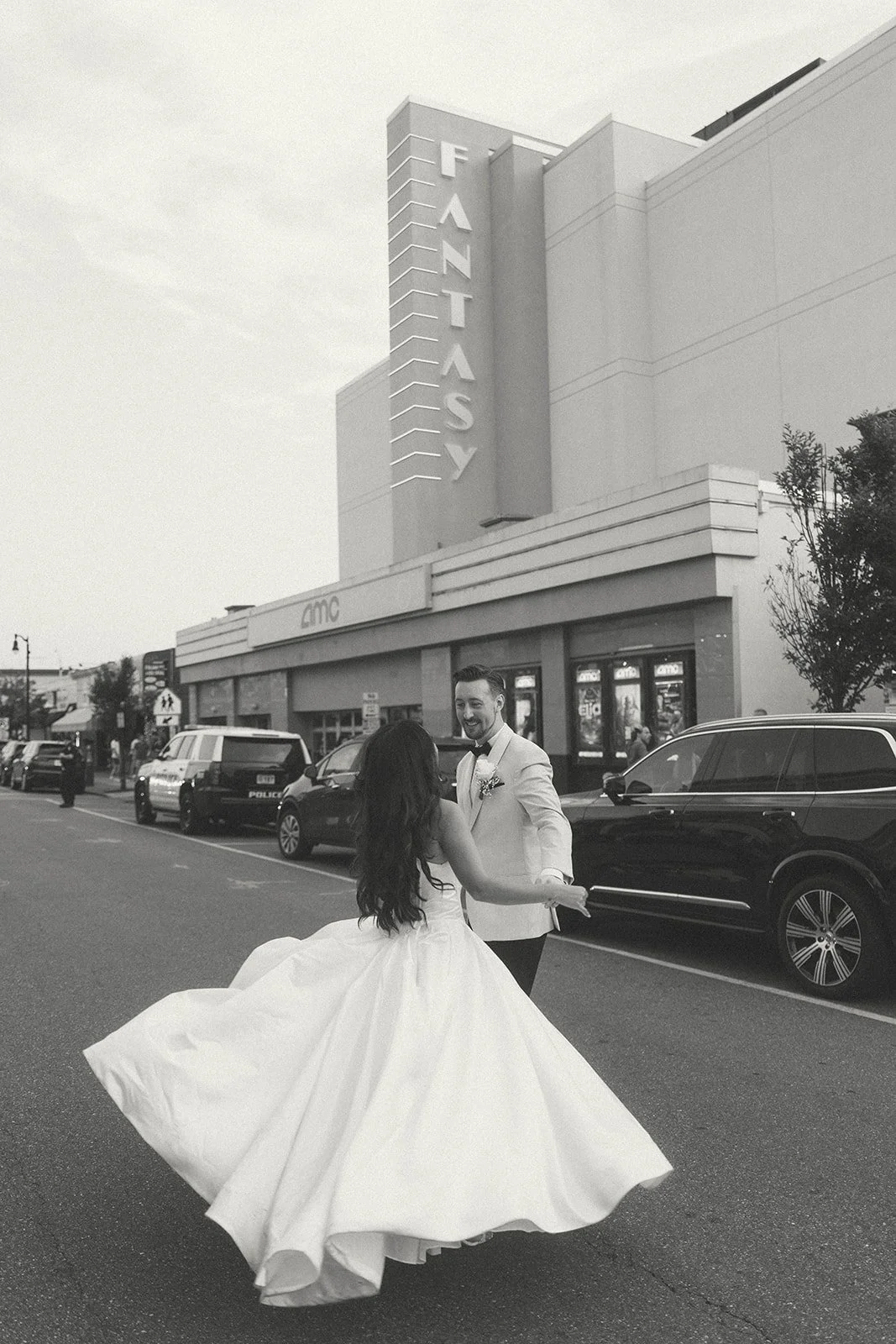 A newlywed couple dancing outside the Fantasy movie theater in front of an AMC building, with cars parked along the street and a few onlookers in the background, in black and white.