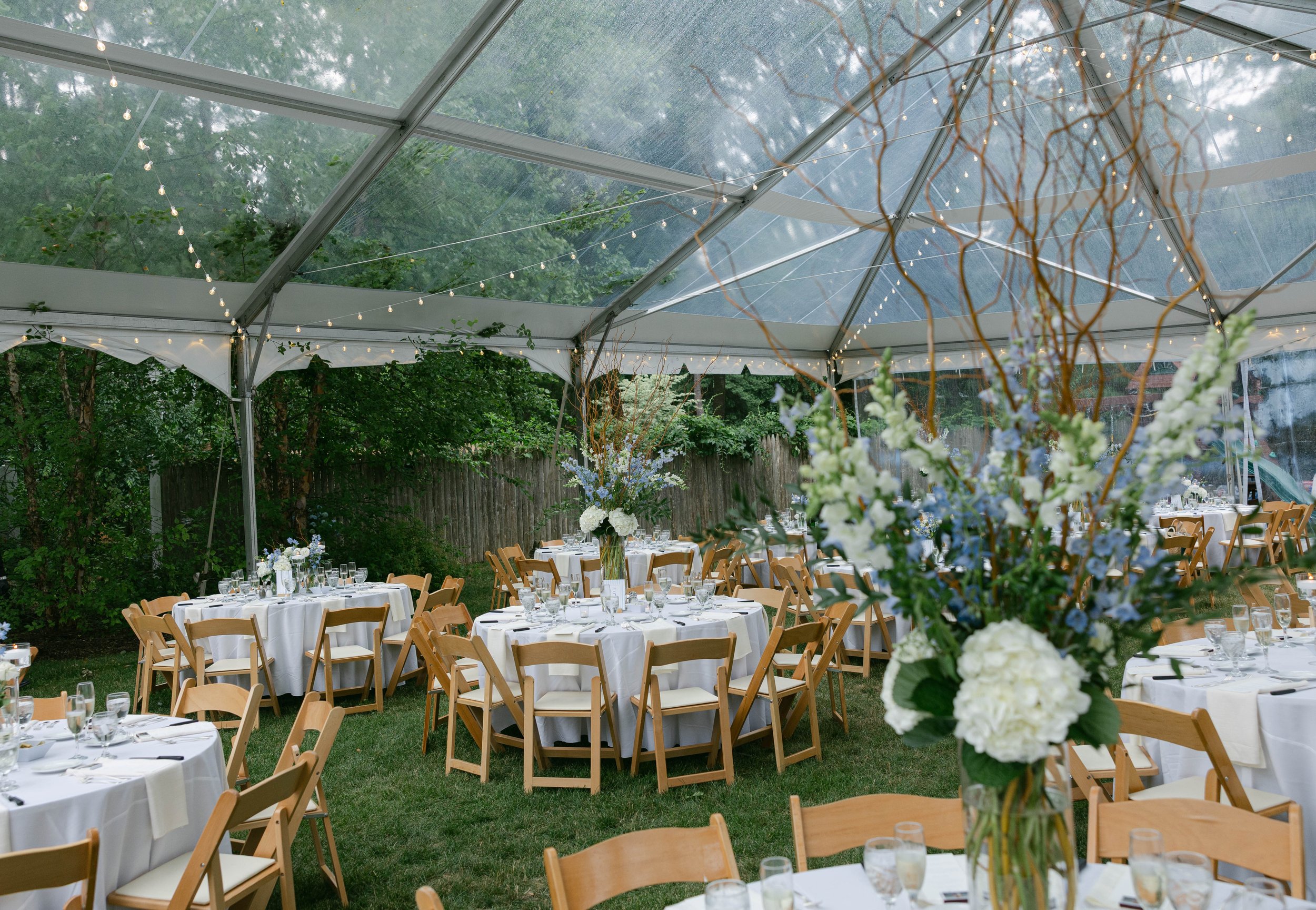 Outdoor event setup with round tables covered in white tablecloths, adorned with centerpieces of white and light blue flowers, and surrounded by wooden chairs. The area is under a large glass tent decorated with string lights, with trees and a fence 
