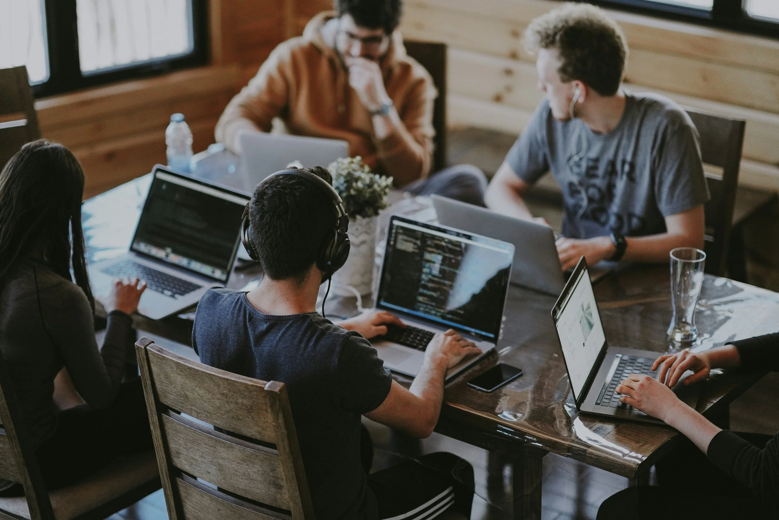 Group of five young adults working on laptops around a wooden dining table in a cozy room with wooden panel walls, some wearing headphones and engaged in their tasks.