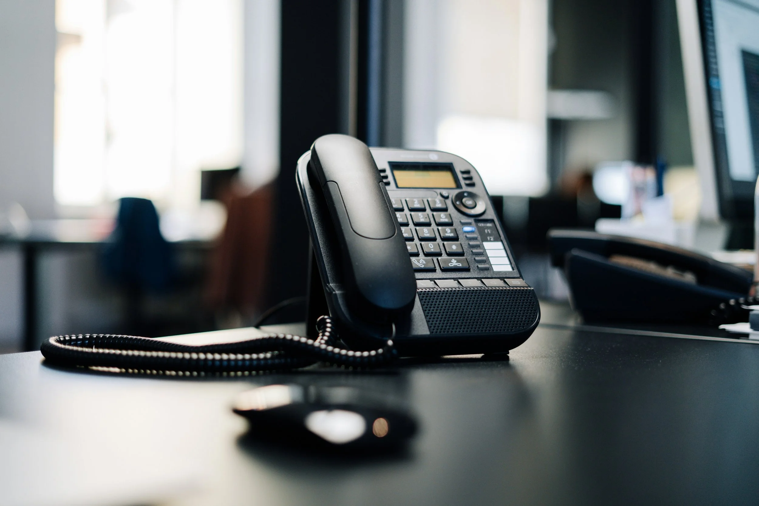 Office desk with a black desktop landline phone, a computer mouse, and a monitor in the background.