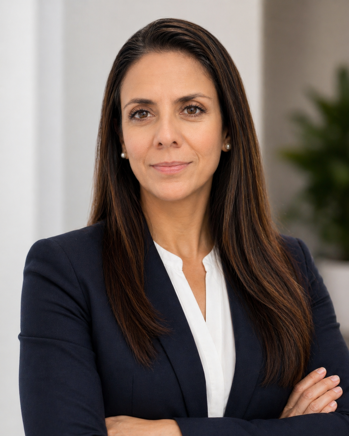 Portrait of a confident woman with long brown hair, wearing a dark blazer and white blouse, standing with arms crossed in a professional setting.