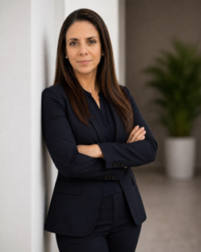 Professional woman in a navy blazer and matching pants with her arms crossed, standing indoors near a white wall with a blurred green plant in the background.