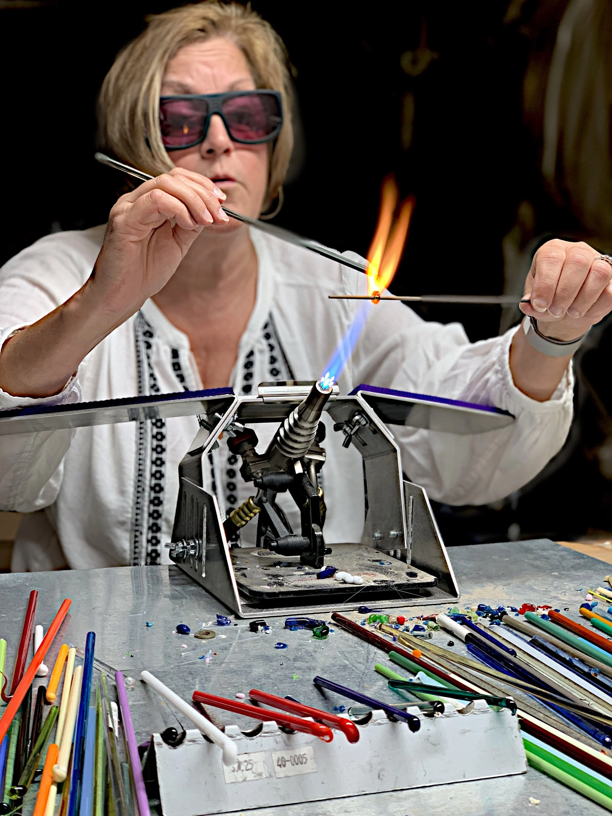 Woman wearing 3D glasses making art with a glassblowing torch, forming glass objects at a workbench with colorful glass rods and tools.