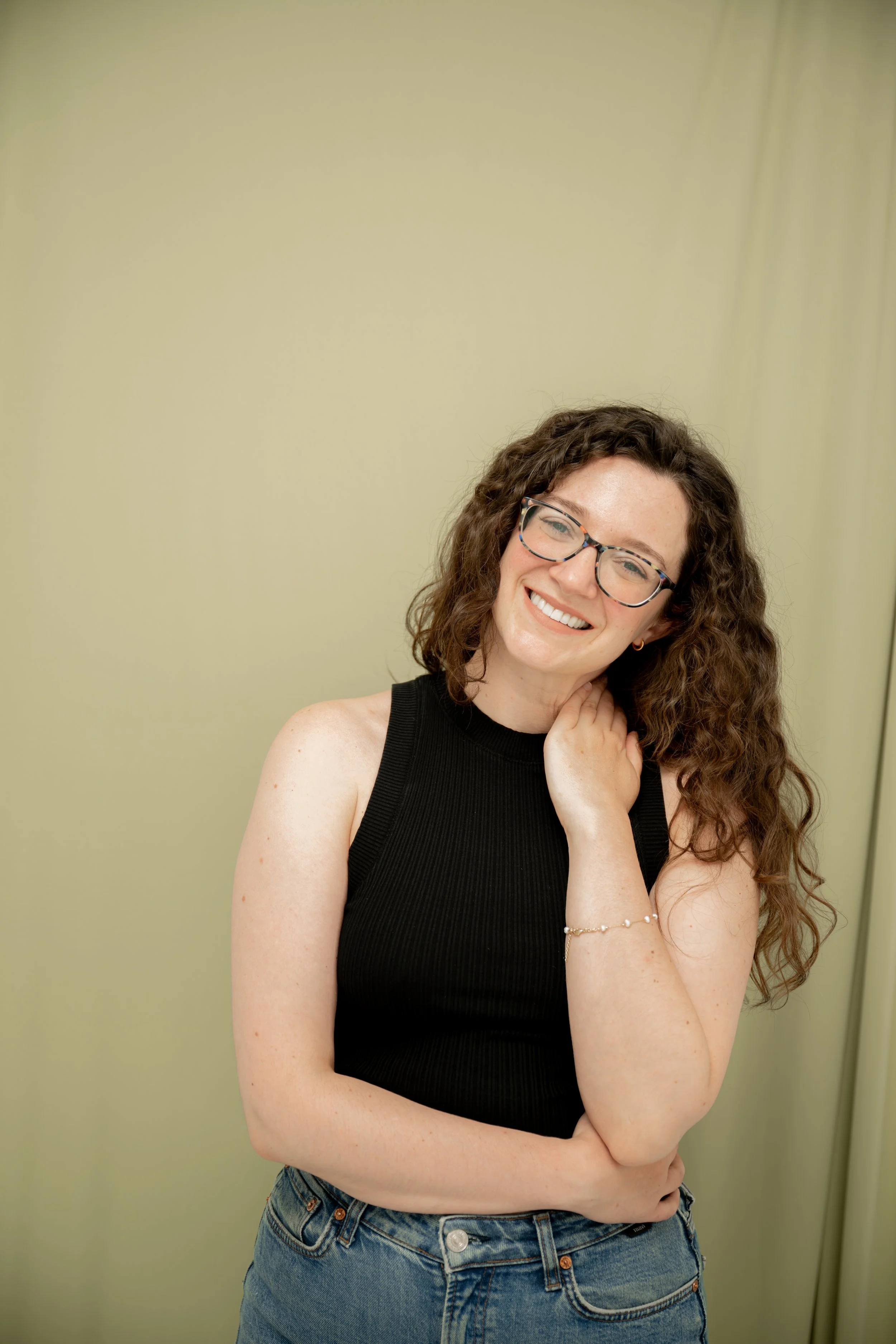 A woman with curly brown hair, glasses, wearing a black sleeveless top and blue jeans, smiling and looking at the camera against a plain, light green wall.