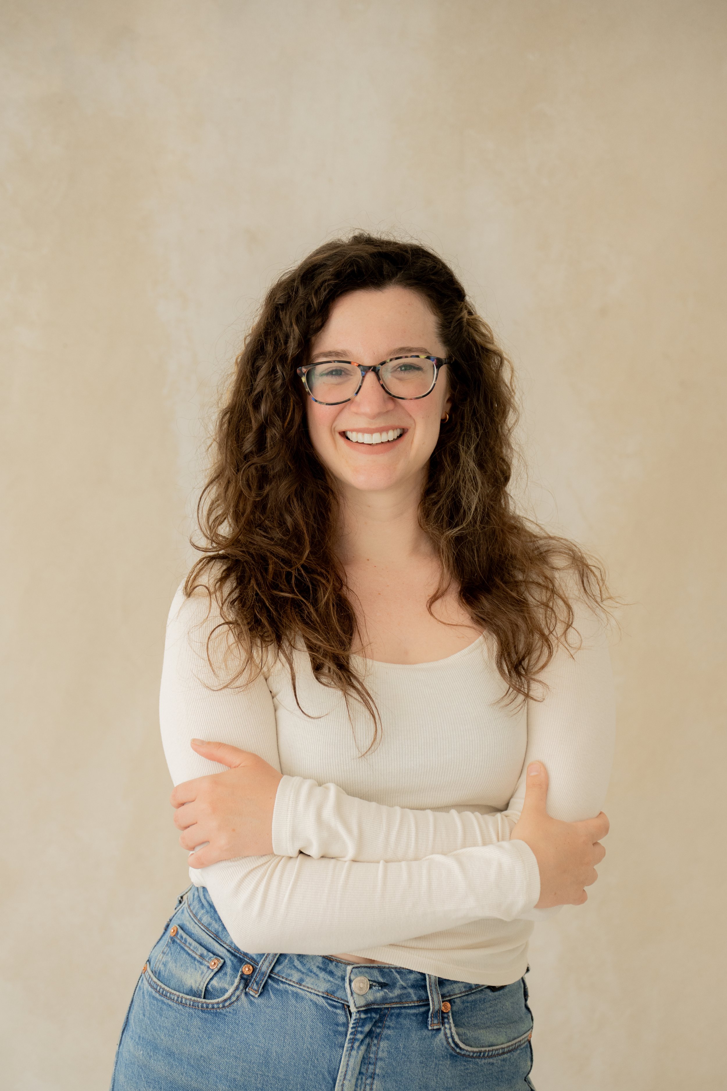 A young woman with curly brown hair, glasses, and a big smile, standing against a plain, beige wall. She is wearing a white long-sleeved top and blue jeans, with her arms crossed.