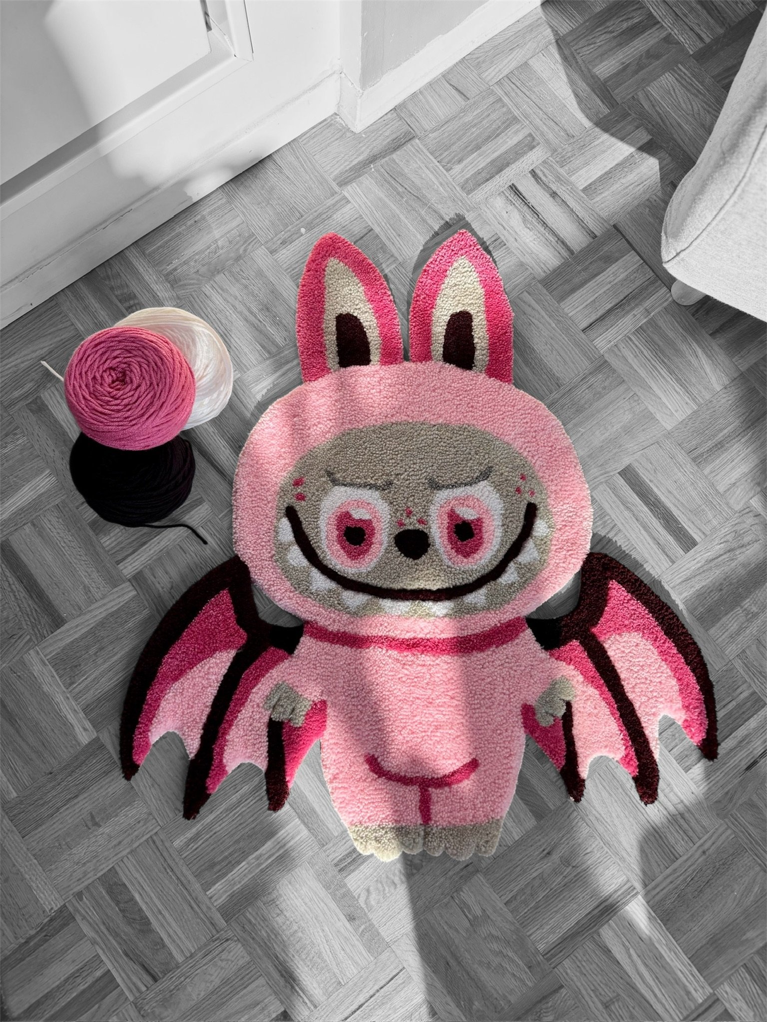Close-up of a pink, rabbit-themed rug on a grey checkered wooden floor, with balls of yarn nearby.