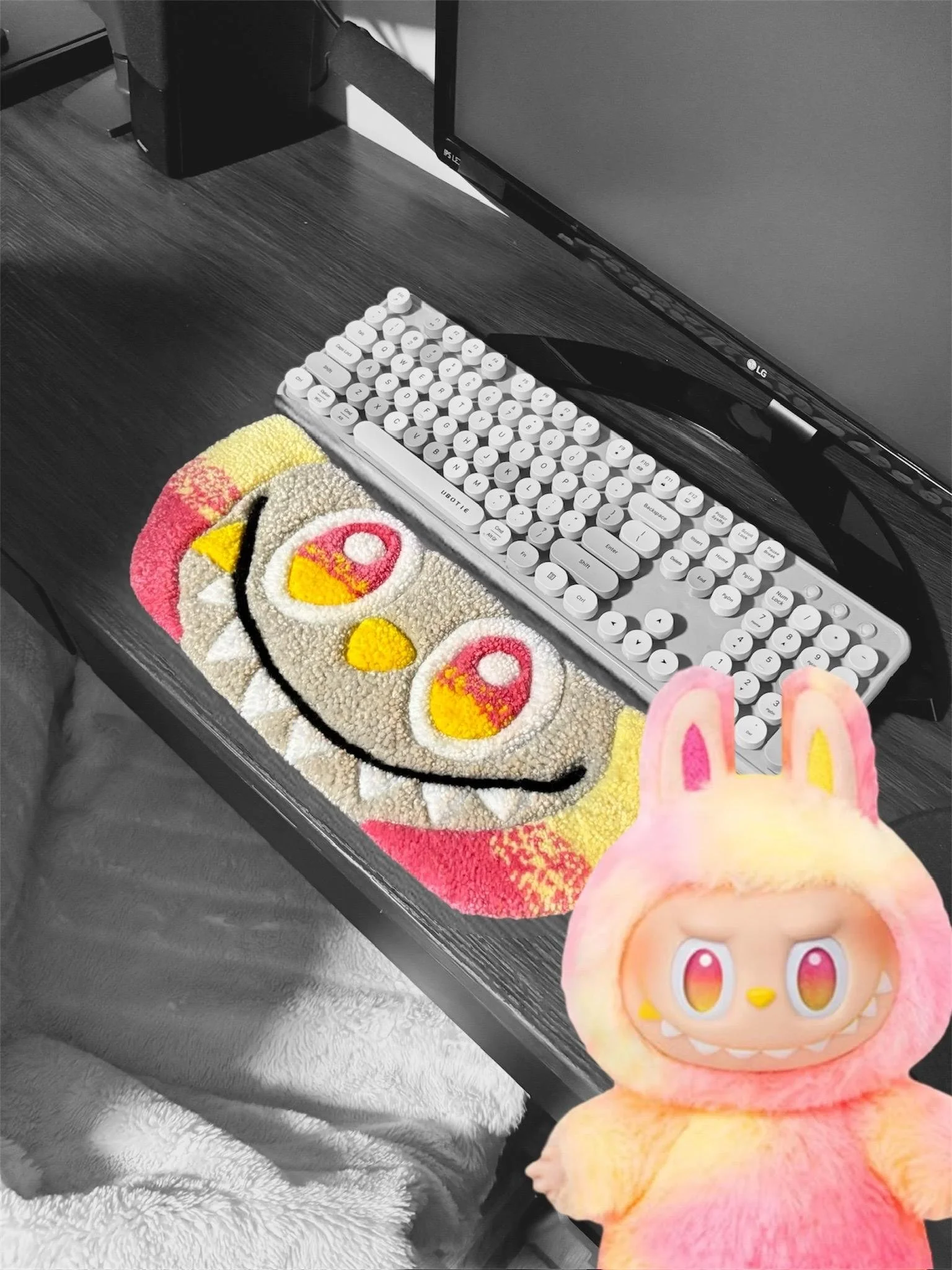 Desk with a computer monitor, a keyboard, a colorful plush mouse pad with cartoon eyes and a smile, and a plush toy with rabbit ears and a monster-like face in vibrant colors.