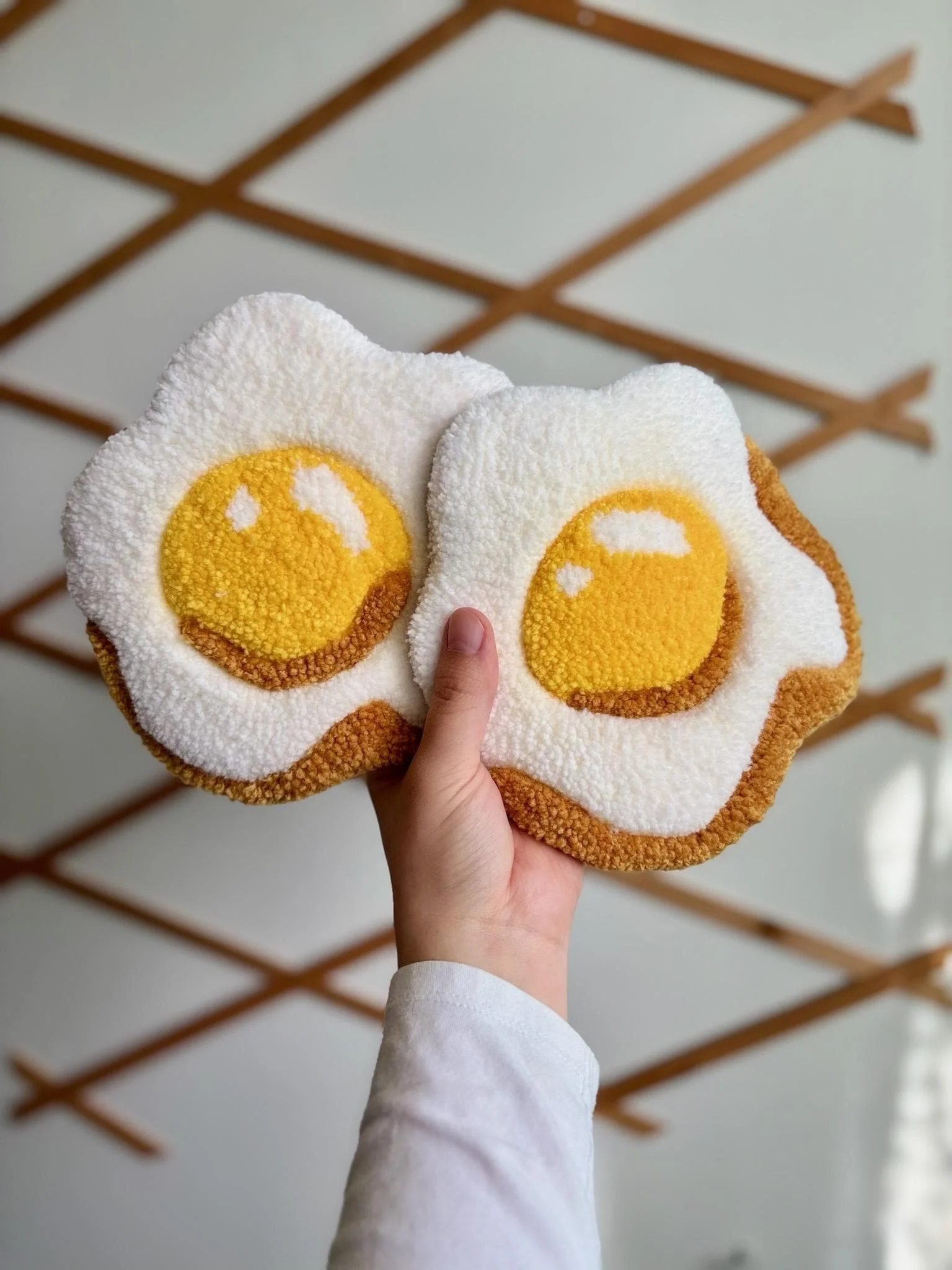 Hand holding two bath mats designed to look like fried eggs with white, yellow, and brown colors, against a background with wooden beams on the ceiling.