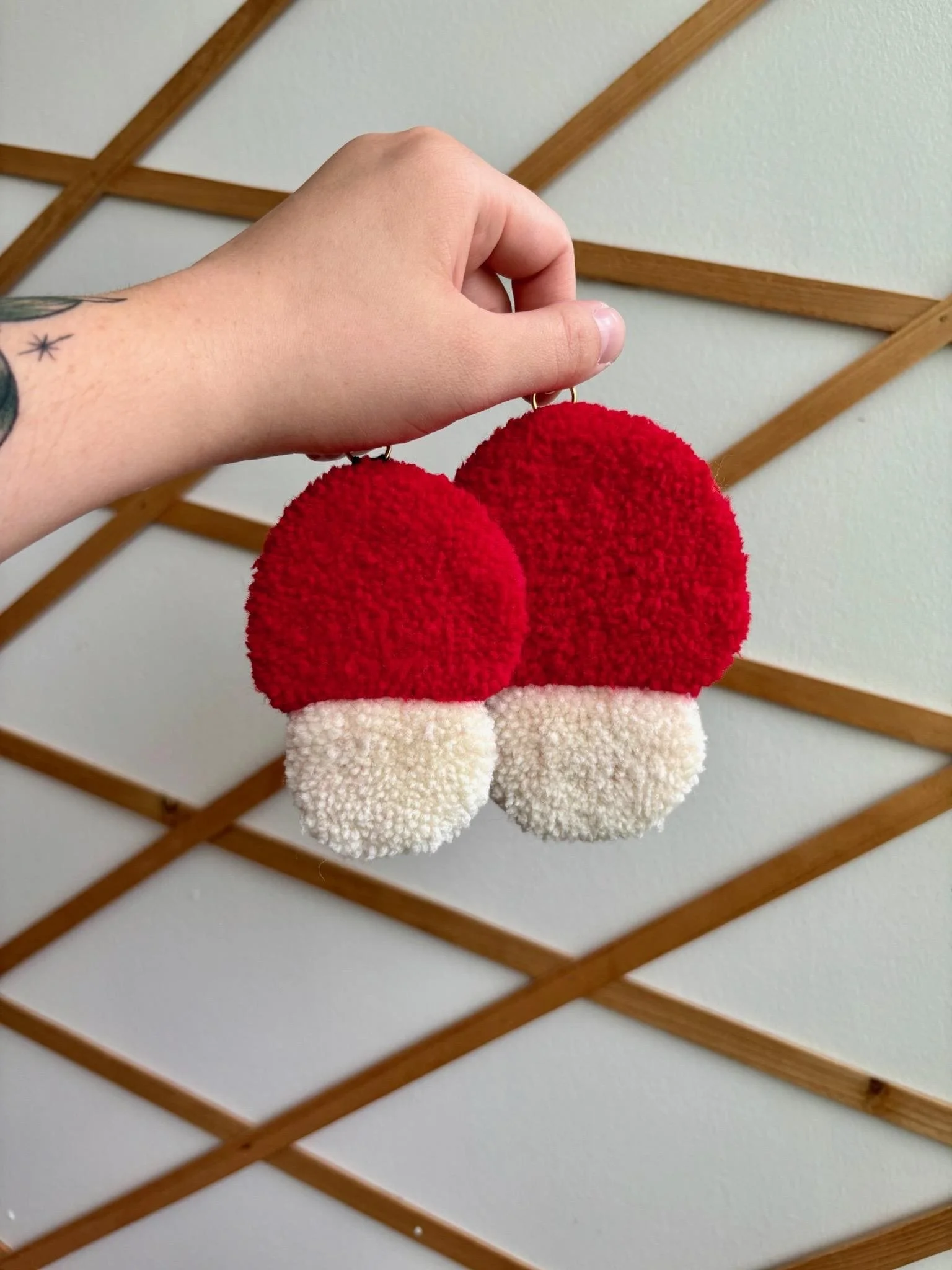 Person holding a pair of red and white fluffy pom-pom earrings in front of a white wall with a wooden lattice.