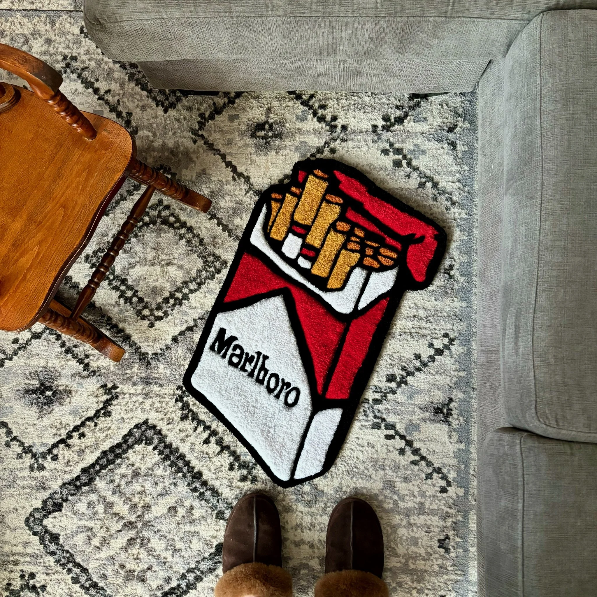 View of a living room floor with a gray patterned area rug, a gray sofa, a wooden chair, and a Wonder Woman themed rug. The photo is taken from a person's perspective, wearing brown shoes and fuzzy socks.