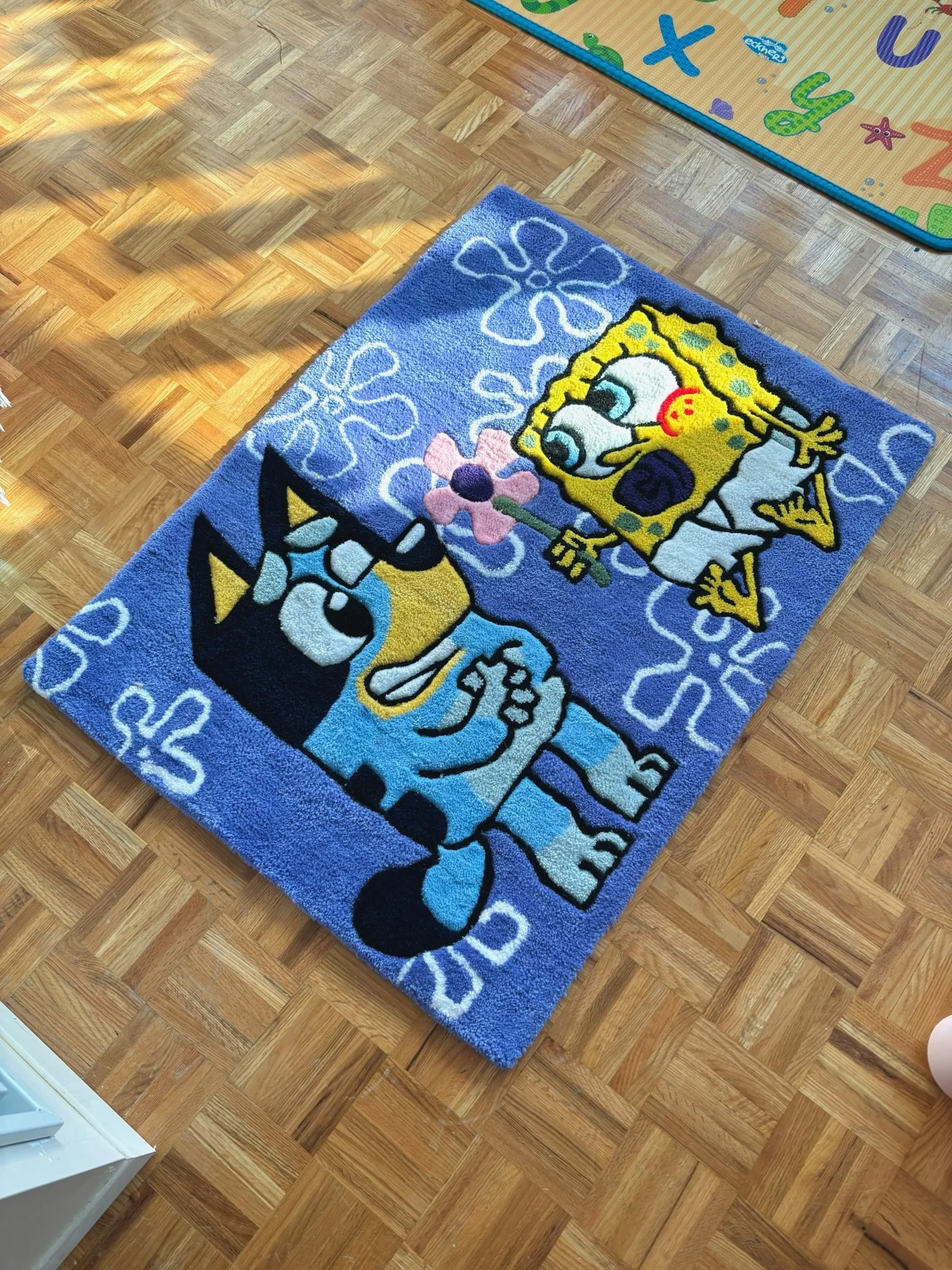 A blue rug featuring SpongeBob SquarePants and Patrick Star from the cartoon, along with flower and bubble patterns, placed on a wooden floor.