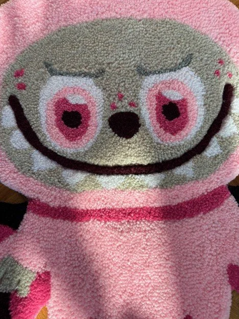 Close-up of a plush toy character with a smiling face, large pink and white eyes, and pink clothing, made from a textured, fluffy material.
