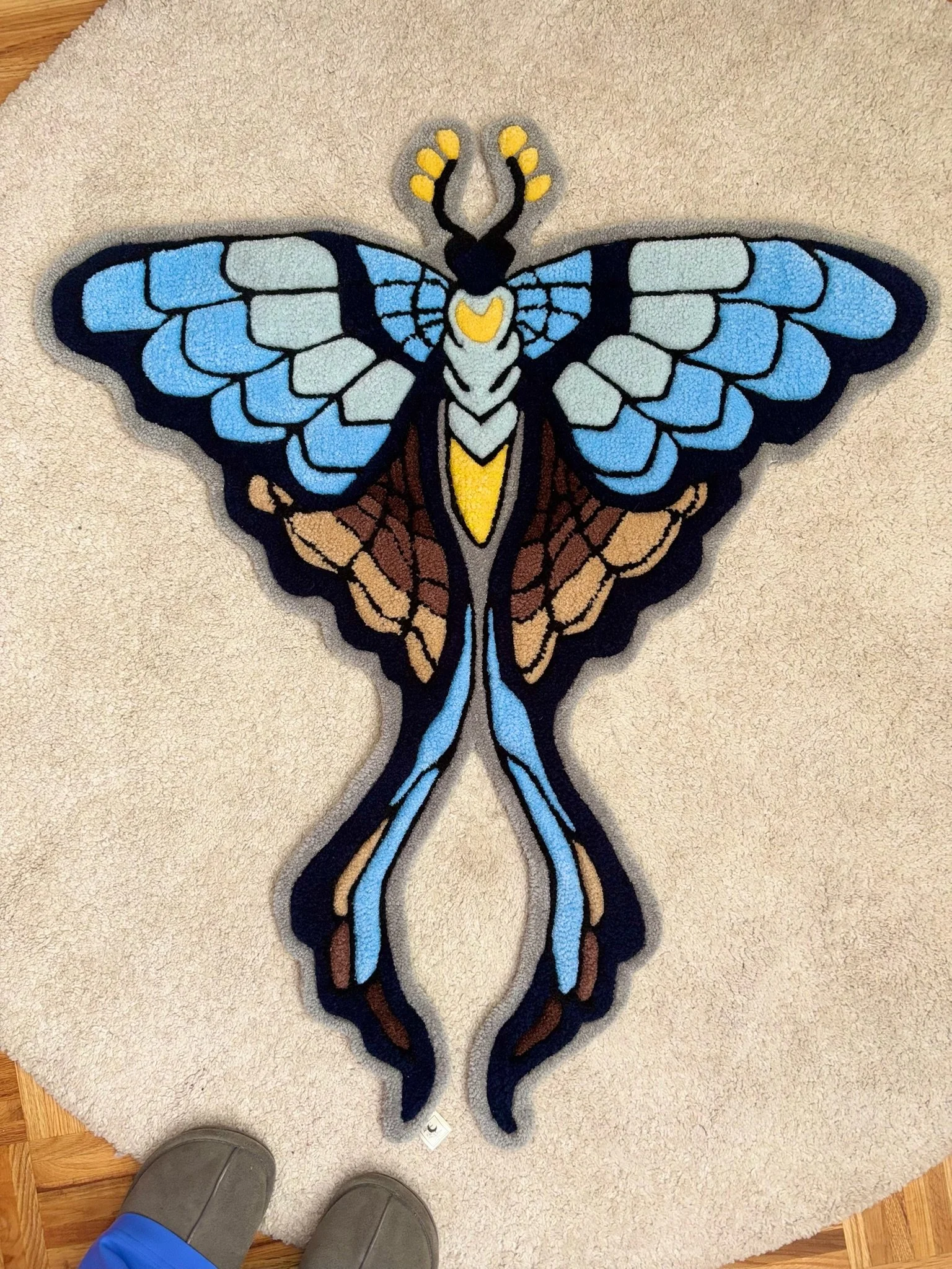 A colorful butterfly-shaped rug on a beige carpet, with the feet of a person wearing gray slippers and blue pants visible at the bottom of the image.