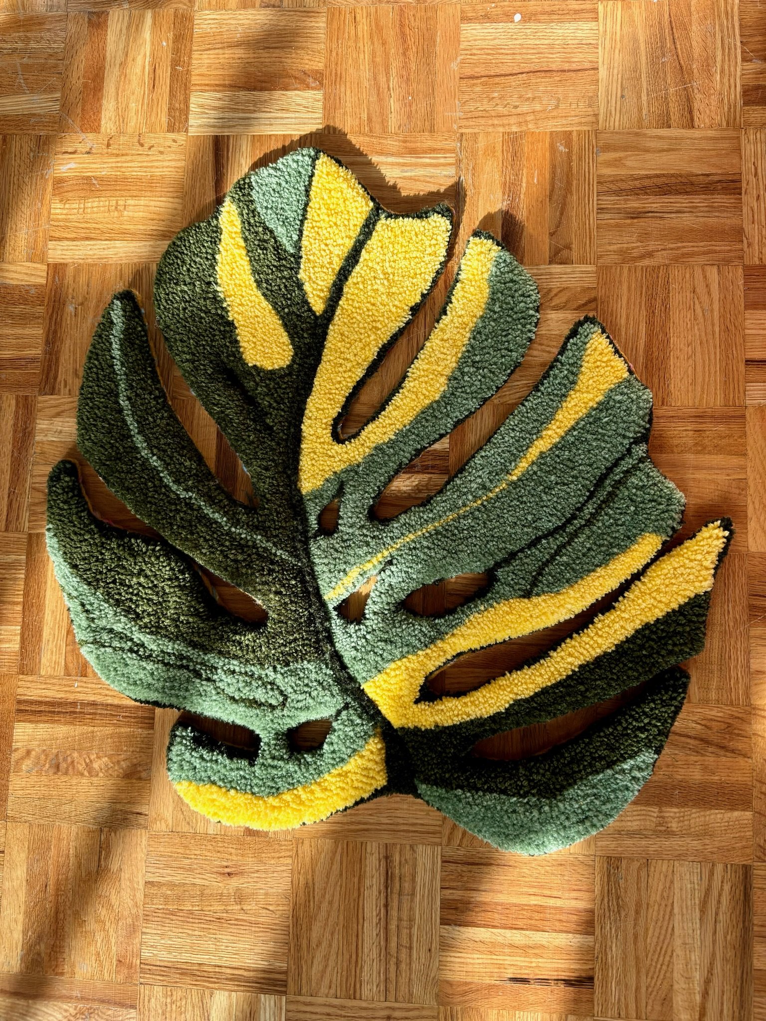 A decorative rug in the shape of a Monstera leaf with green and yellow colors resting on a wooden parquet floor.
