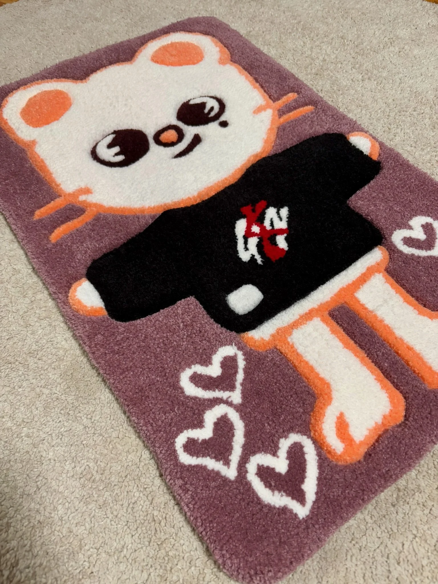 A pink rectangular rug with a cartoon hamster holding a smartphone, surrounded by white hearts, on a beige carpet.