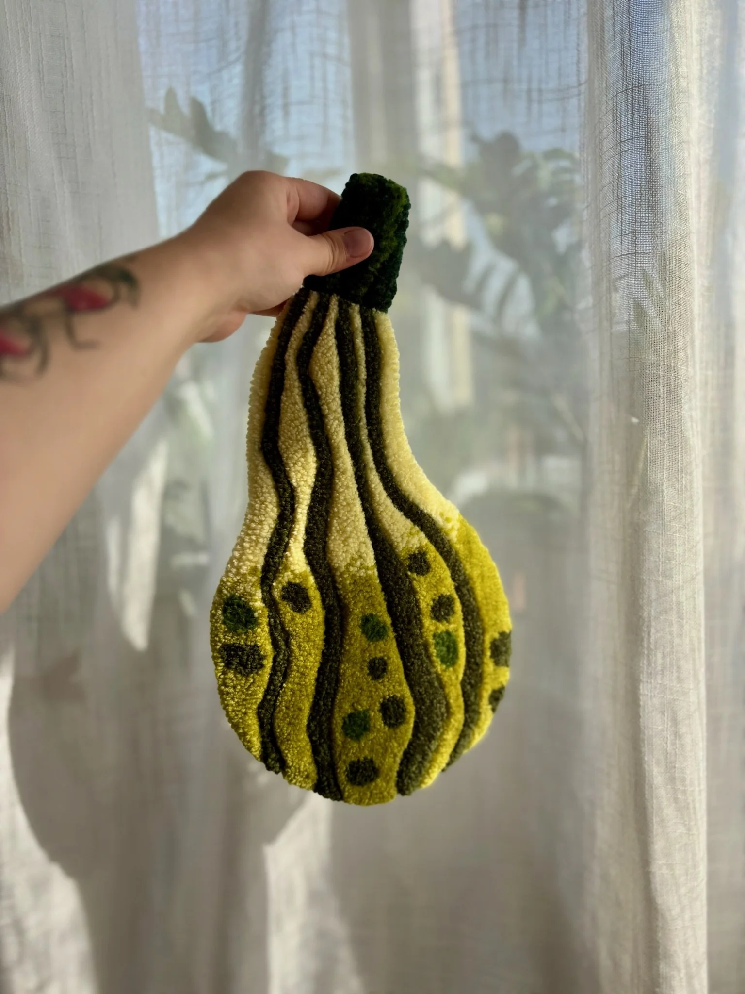 A hand holding a yellow and black zebra-striped cleaning glove in front of a semi-transparent curtain with plants visible behind it.