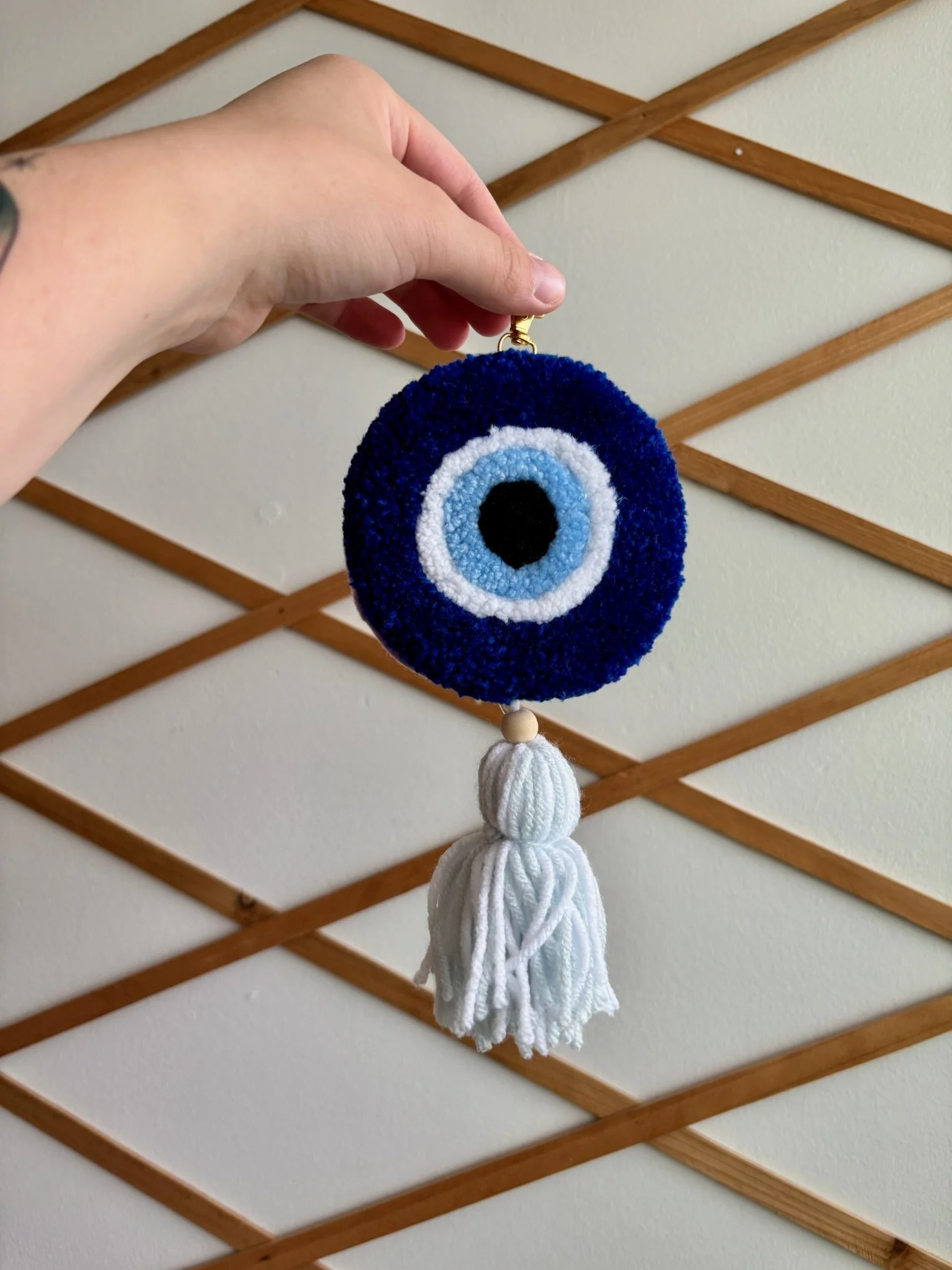 Hand holding a circular felt ornament with a blue and black eye design, hanging from a wooden lattice wall.
