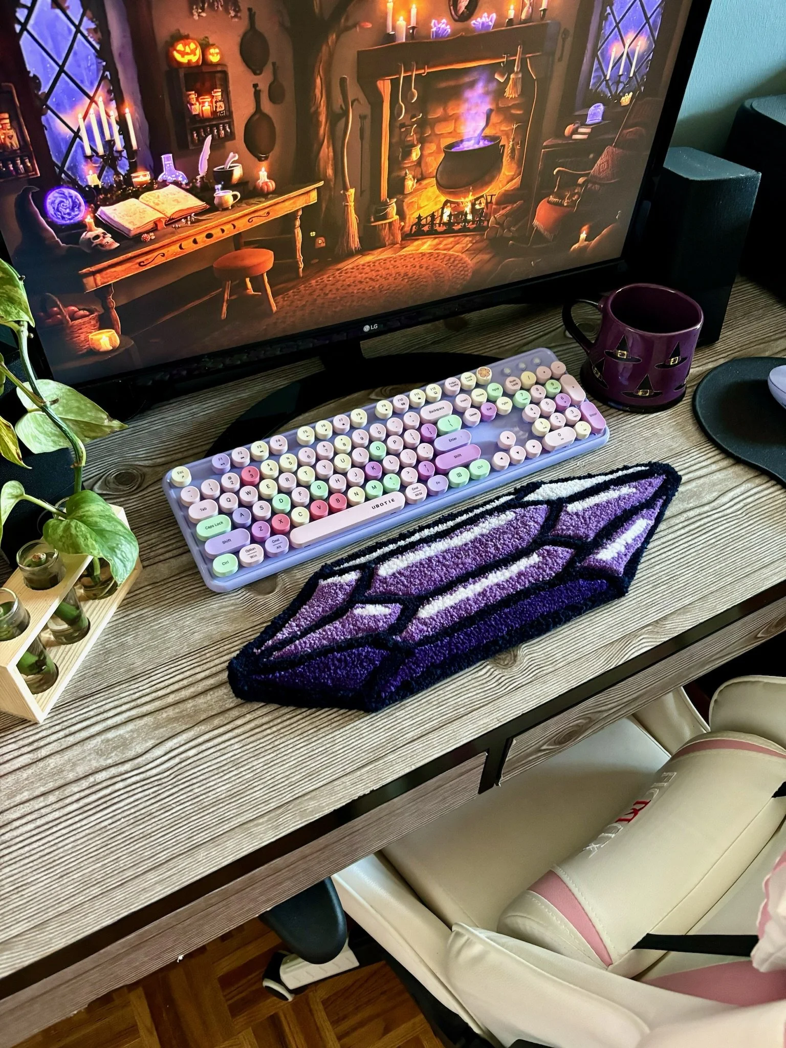 The image shows a computer monitor on a wooden desk displaying a spooky Halloween-themed scene with a fireplace, candles, and Halloween decorations. In front of the monitor is a pastel-colored mechanical keyboard, and to the right is a purple Halloween mug. There is a black mouse pad with a mouse, and a plant with green leaves is on the left side of the desk. Under the desk, there is a cream-colored chair with pink accents.