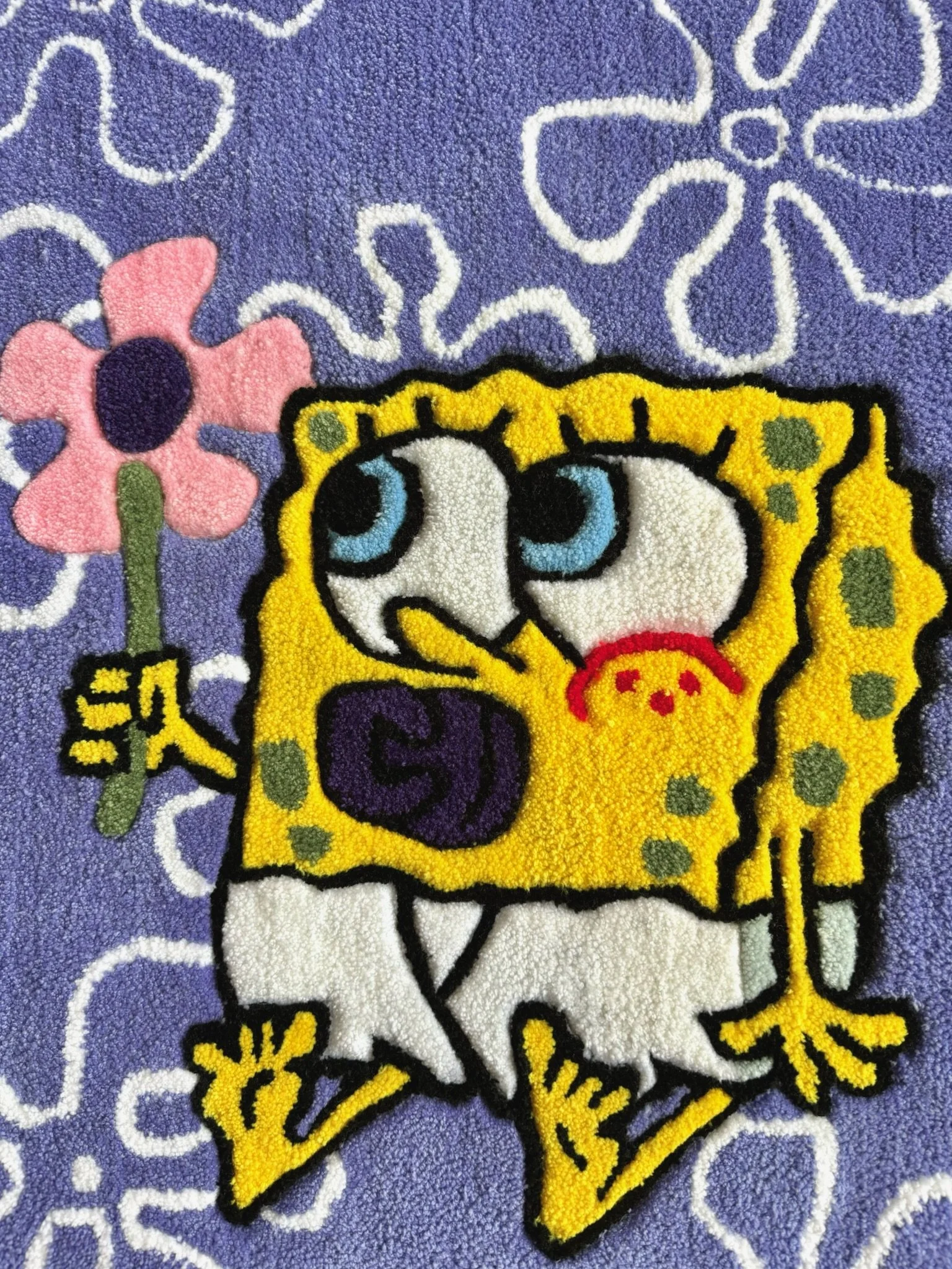 A colorful cartoon design on a textured purple background features SpongeBob SquarePants with large blue eyes, a big nose, and a wide smile. He is holding a pink flower with a black center in his left hand, and has a small red crab on his chest near his mouth.