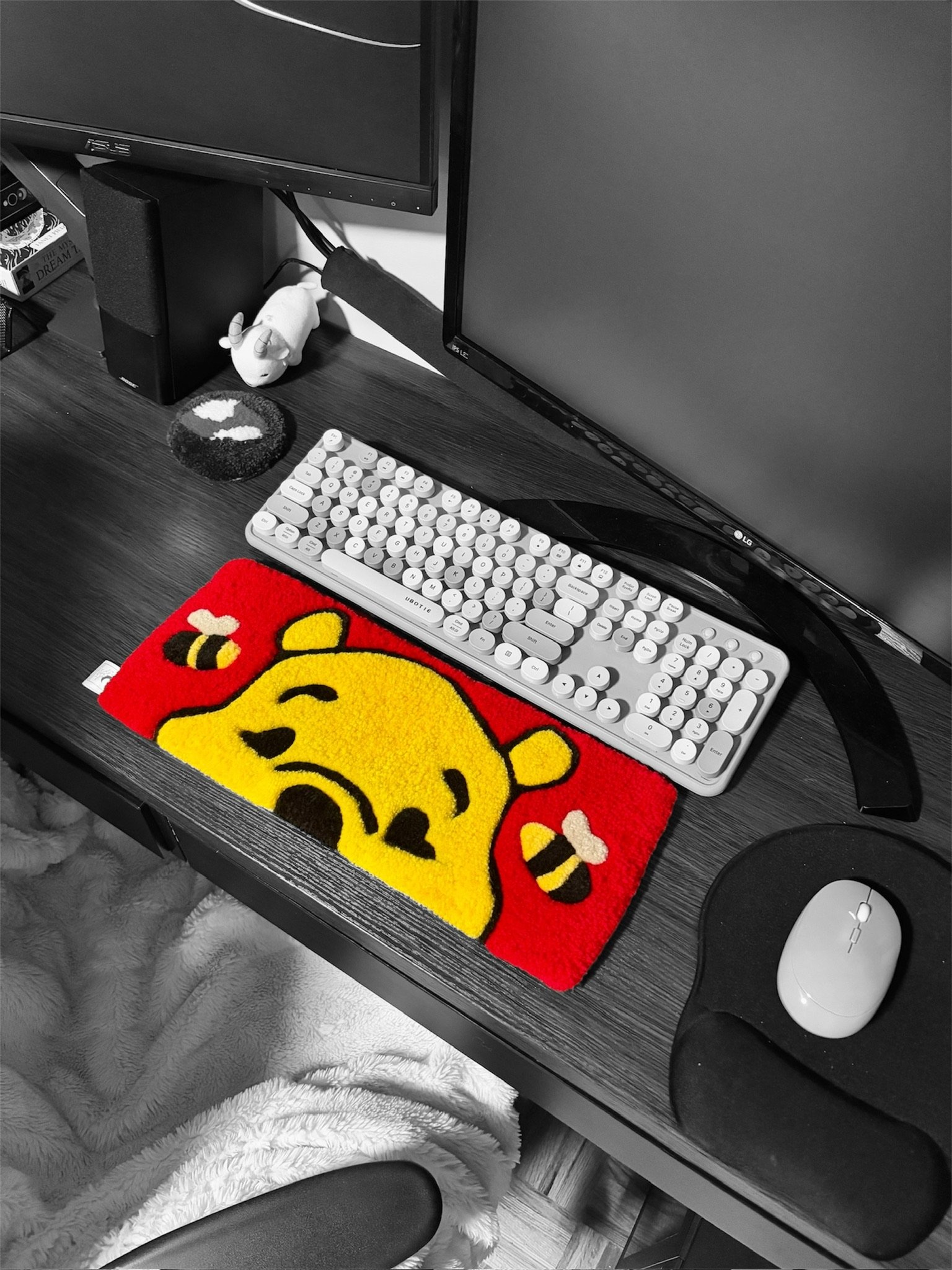 A computer desk with a monitor, keyboard, mouse, and a colorful plush mat with a cartoon character on it. There is a small plush toy mouse on the desk and a speaker next to the monitor. A fluffy blanket is draped over a chair under the desk.