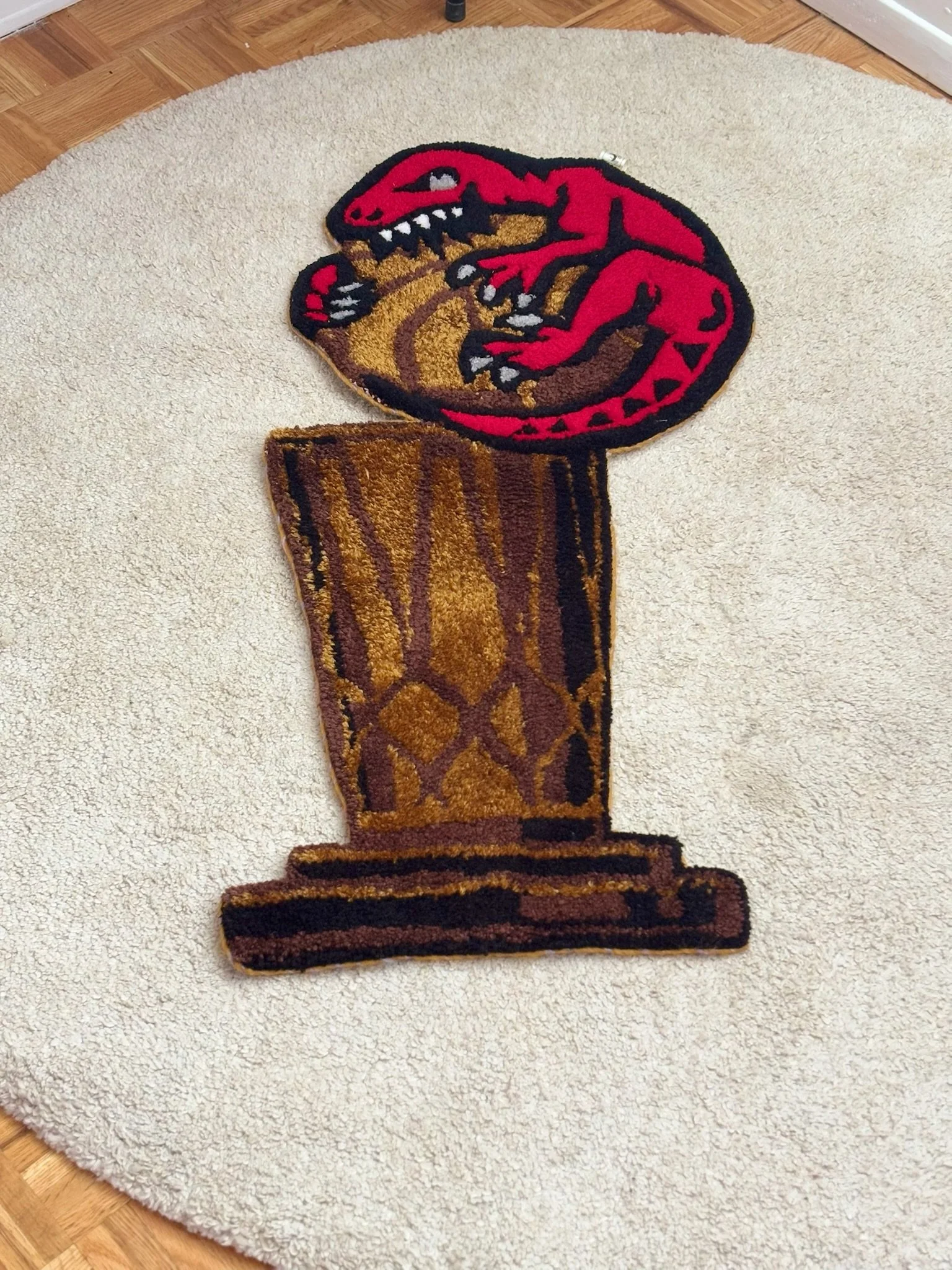A colorful rug shaped like a T-Rex dinosaur holding a goblet, placed on a white round rug on a wooden floor.