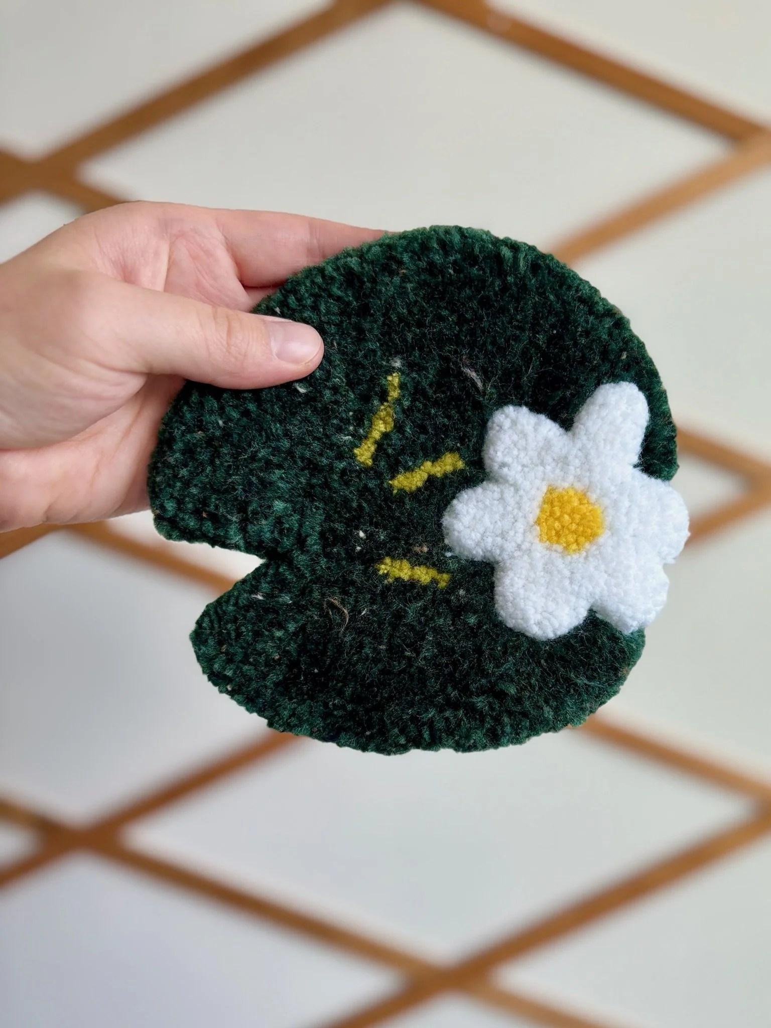Close-up of a person's hand holding a dark green fuzzy scrubbing pad with a white and yellow flower decoration attached to it, blurred background with a wooden lattice.