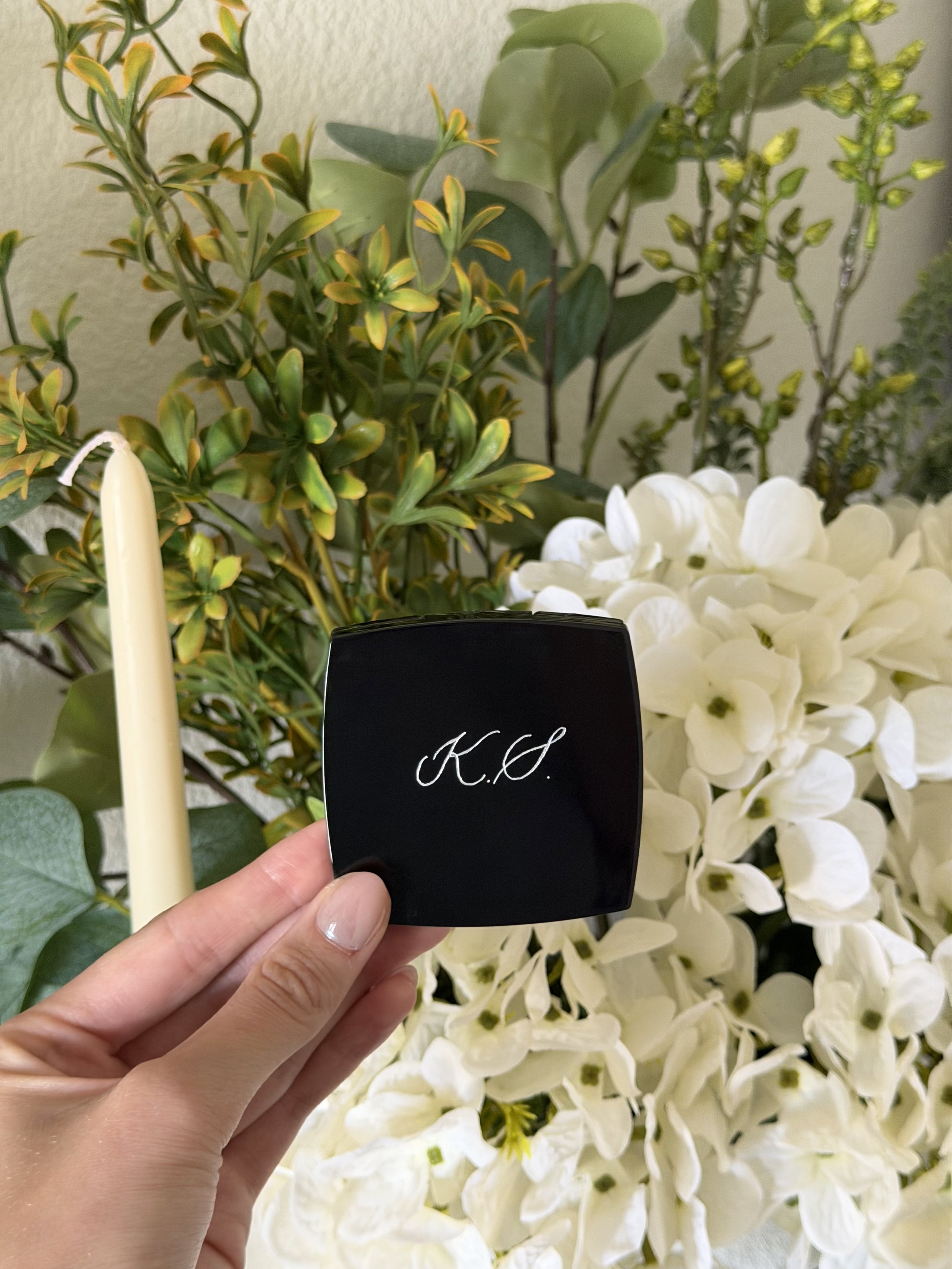 Black compact mirror personalized with custom initials, photographed with white flowers.