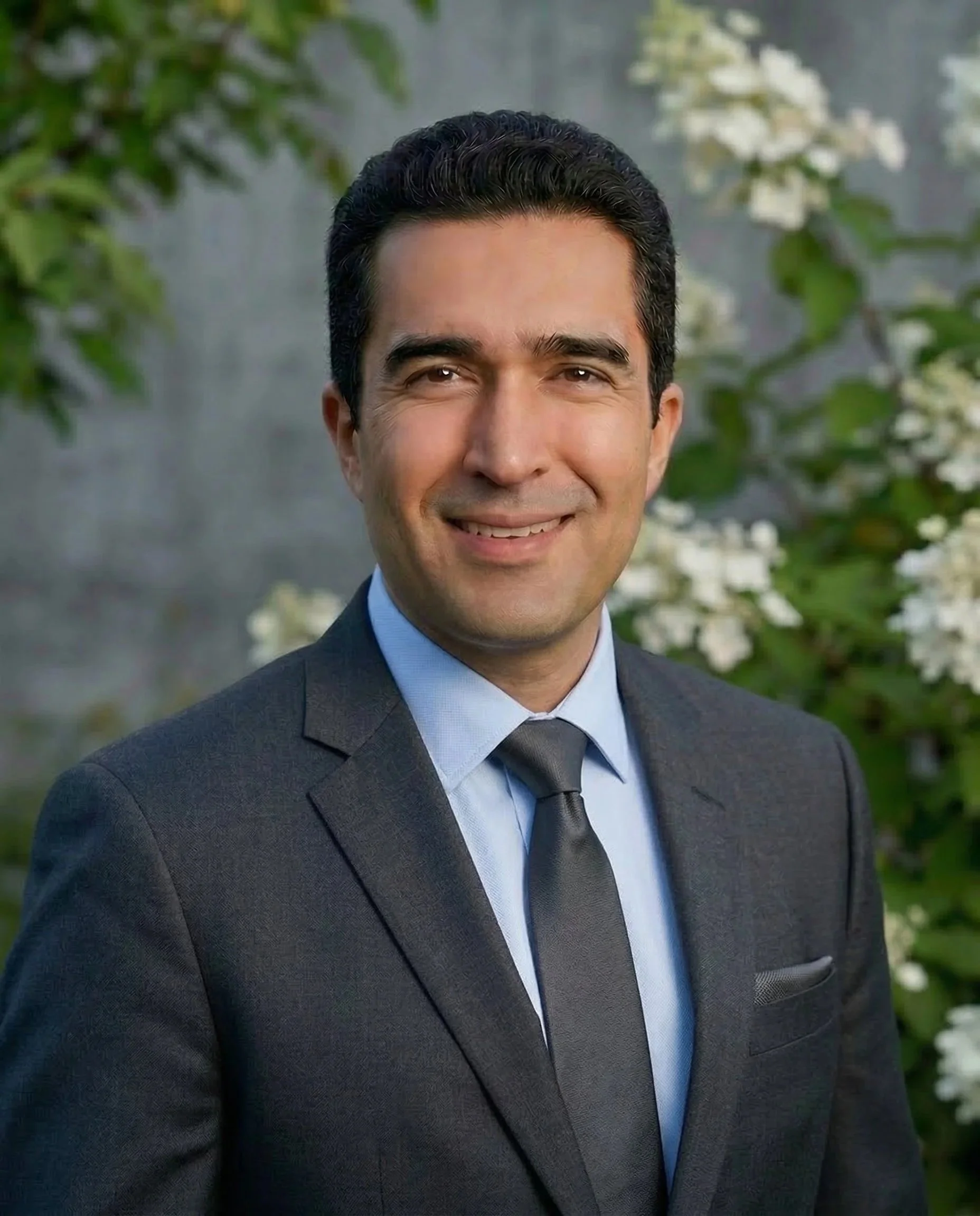 A man in a dark suit with a light blue shirt and black tie, smiling outdoors with greenery and white flowers in the background.