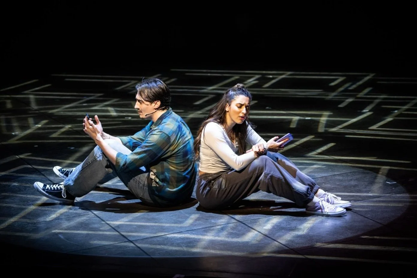 Two actors sitting back to back on a dark stage, each focused on a phone, with a geometric light pattern on the floor.