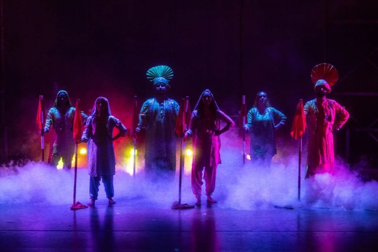 A group of six performers on stage dressed in traditional Indian costumes, standing in a line with colorful flags, with dramatic lighting and fog creating a vibrant atmosphere.