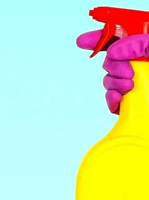 A hand wearing a purple glove holding a yellow spray bottle with a red nozzle against a light blue background.